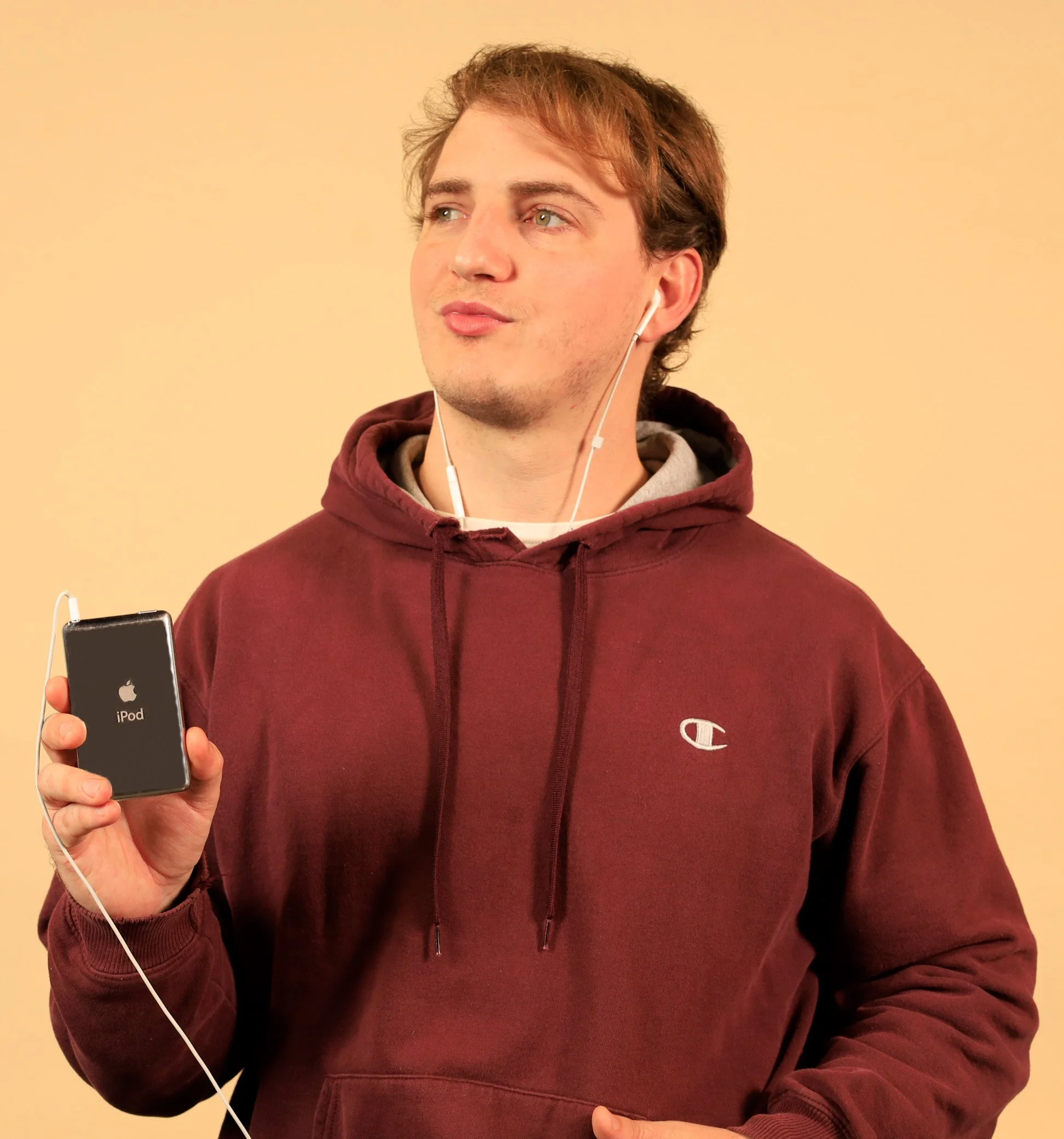 A young man with light-colored hair wearing a maroon hoodie with a gray shirt underneath, listening to white earphones, holding a black iPod, against a plain beige background.