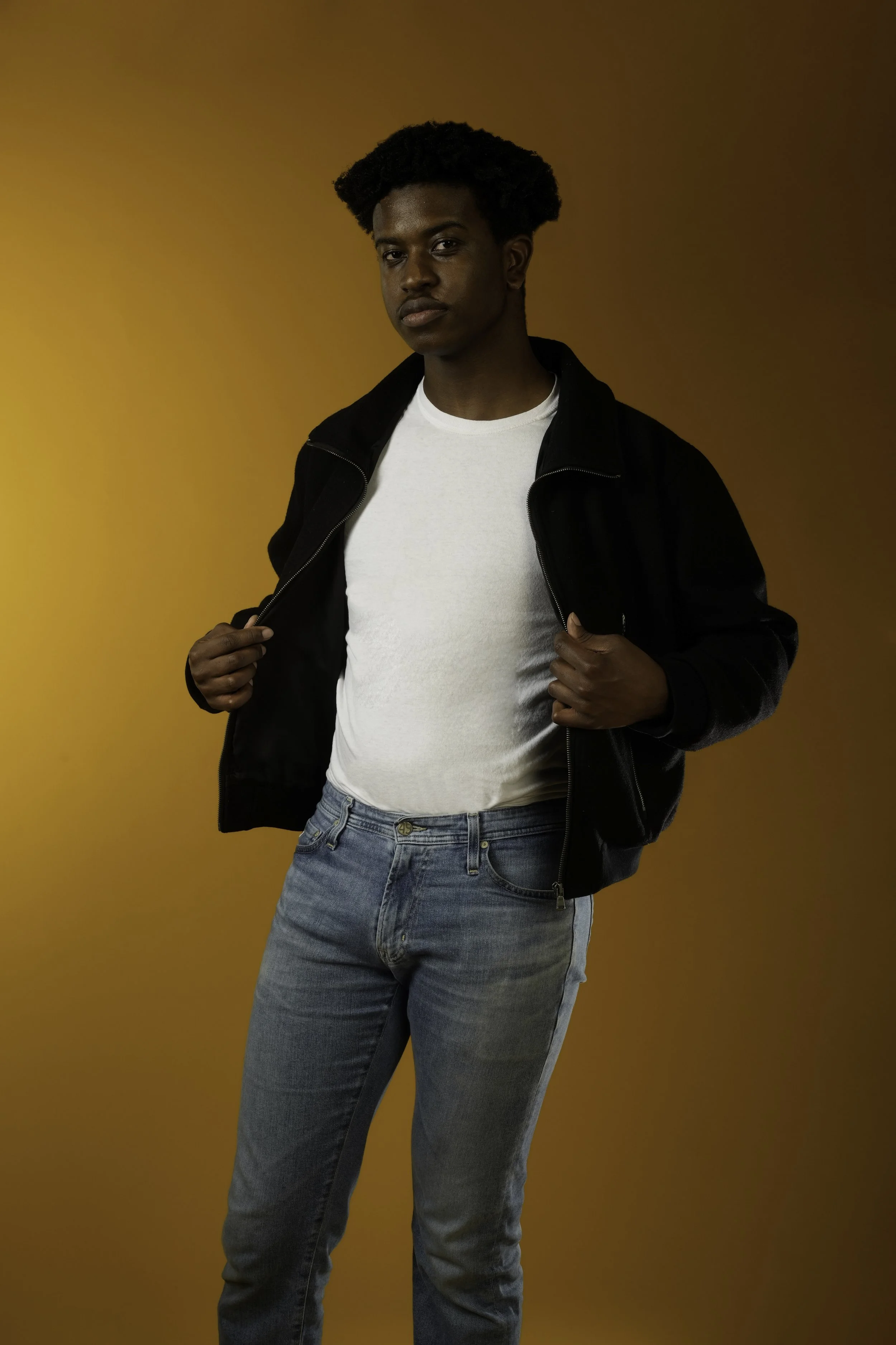 A young man with dark skin and styled afro hair wearing a black jacket over a white T-shirt and light blue jeans, standing against a solid orange background, looking at the camera with a neutral expression, holding the edges of his jacket.