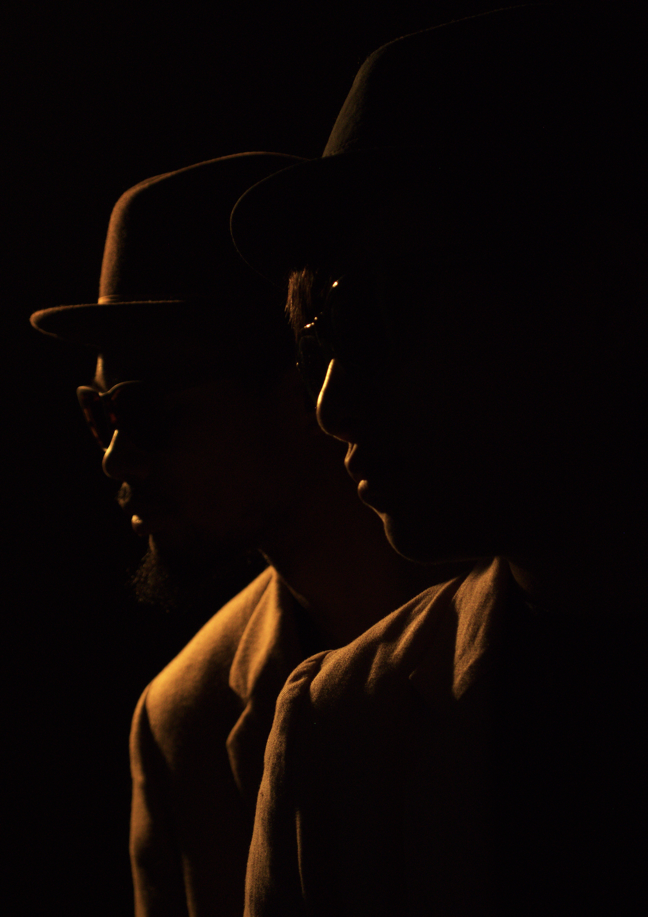 Silhouettes of two people wearing hats and glasses, facing each other in profile, illuminated by a warm, low light source against a dark background.