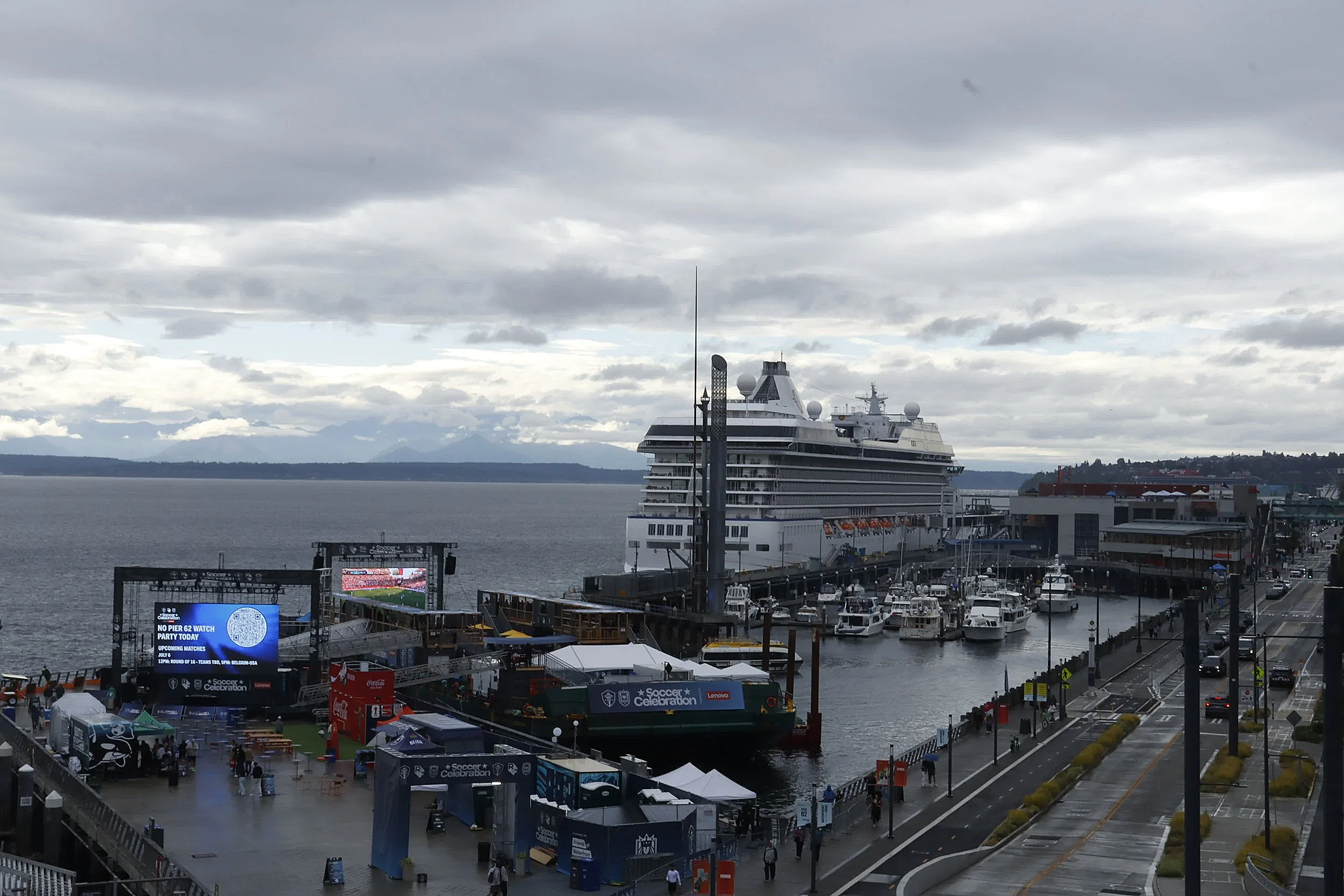 A view of a harbor with a large cruise ship docked next to a pier. The cityscape and mountains are visible in the distance under a cloudy sky. There are boats in the water and various people walking around a busy area near the docks.