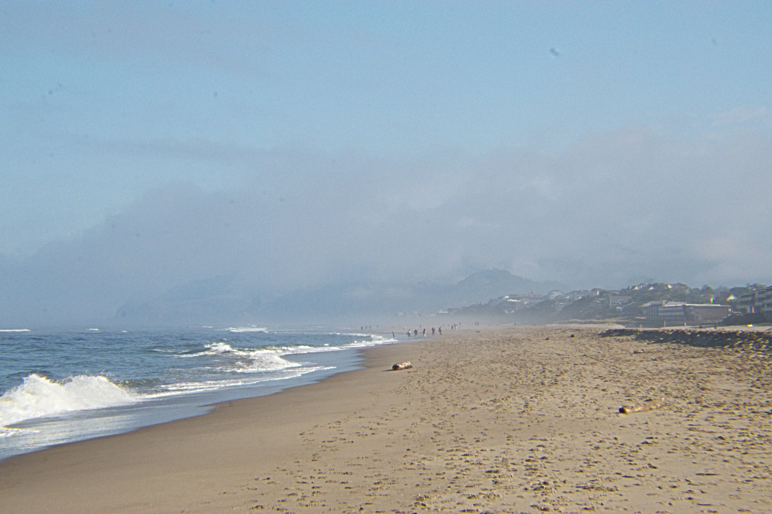 A beach scene with ocean waves, sandy shore, a few people walking, and cliffs or hills in the background under cloudy sky.