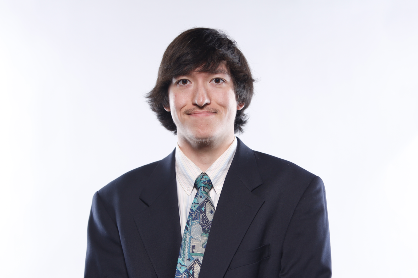 A young man with medium-length dark hair, wearing a dark suit, a white striped shirt, and a colorful patterned tie, standing against a plain white background.