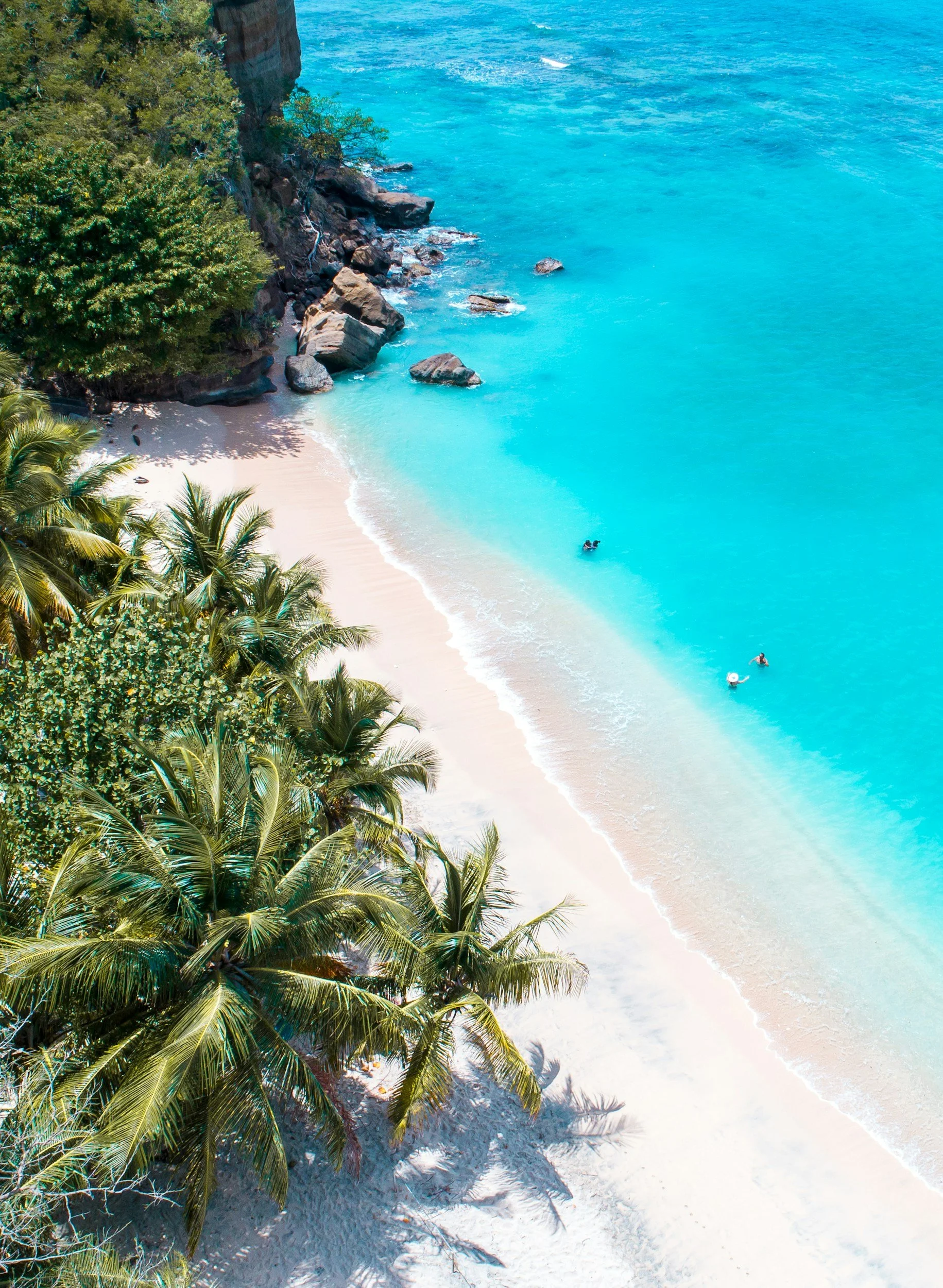 Aerial view of a tropical beach with white sand, turquoise water, lush green palm trees, and rocks along the shoreline.