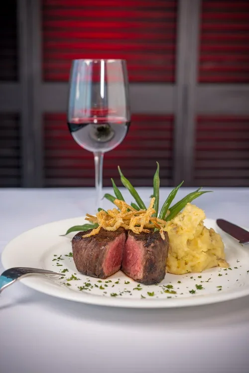 A plate with a cooked steak, green beans, mashed potatoes, and fried onion strings, with a glass of red wine in the background.