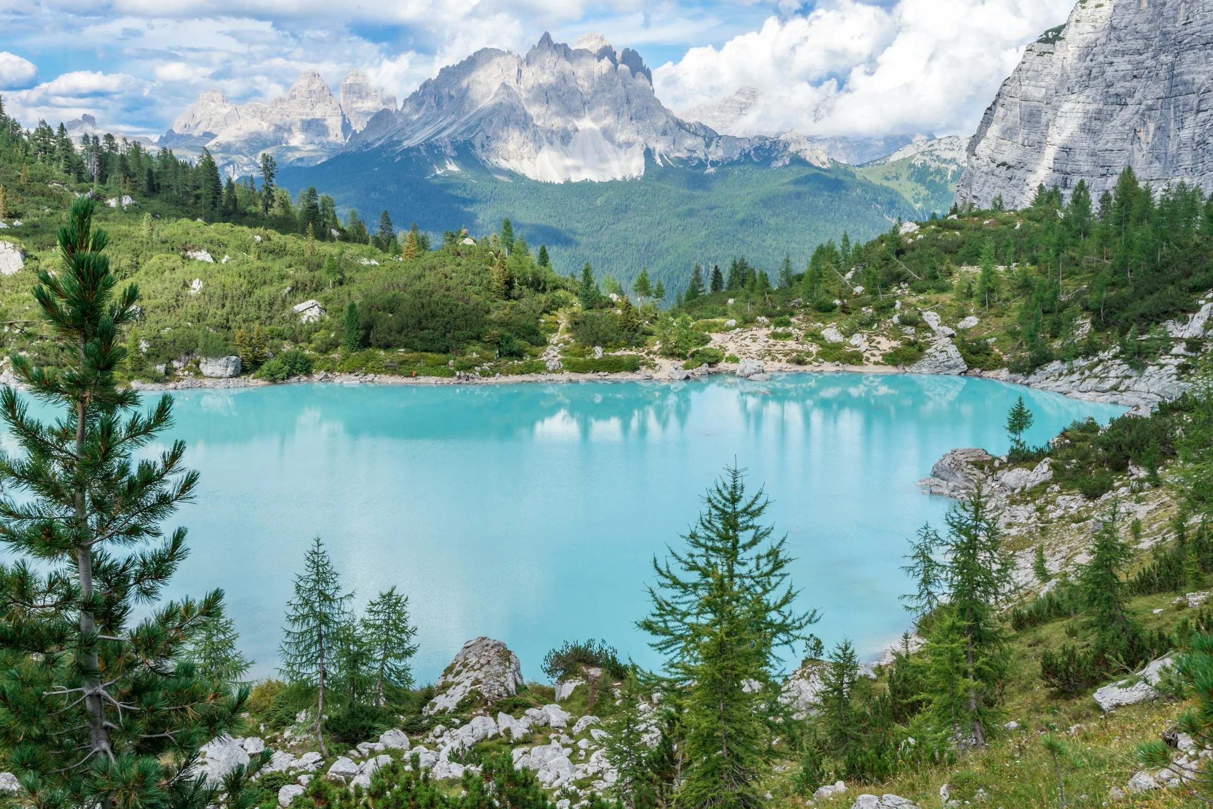 Scenic view of a turquoise alpine lake surrounded by rocky terrain and green pine trees, with tall rugged mountains in the background under a partly cloudy sky.