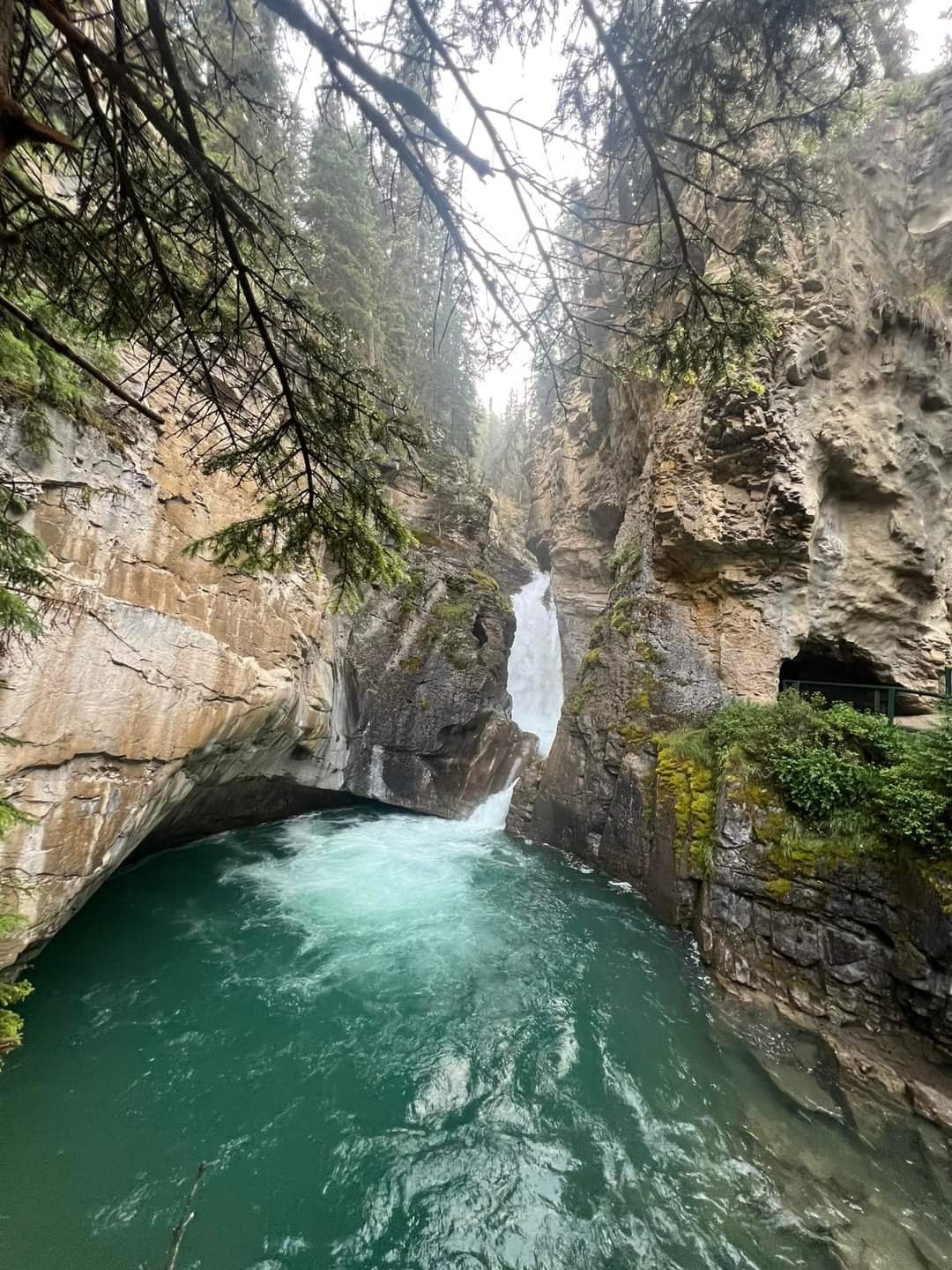 A scenic view of a narrow canyon with a waterfall flowing into a green river, surrounded by rocky cliffs and pine trees.