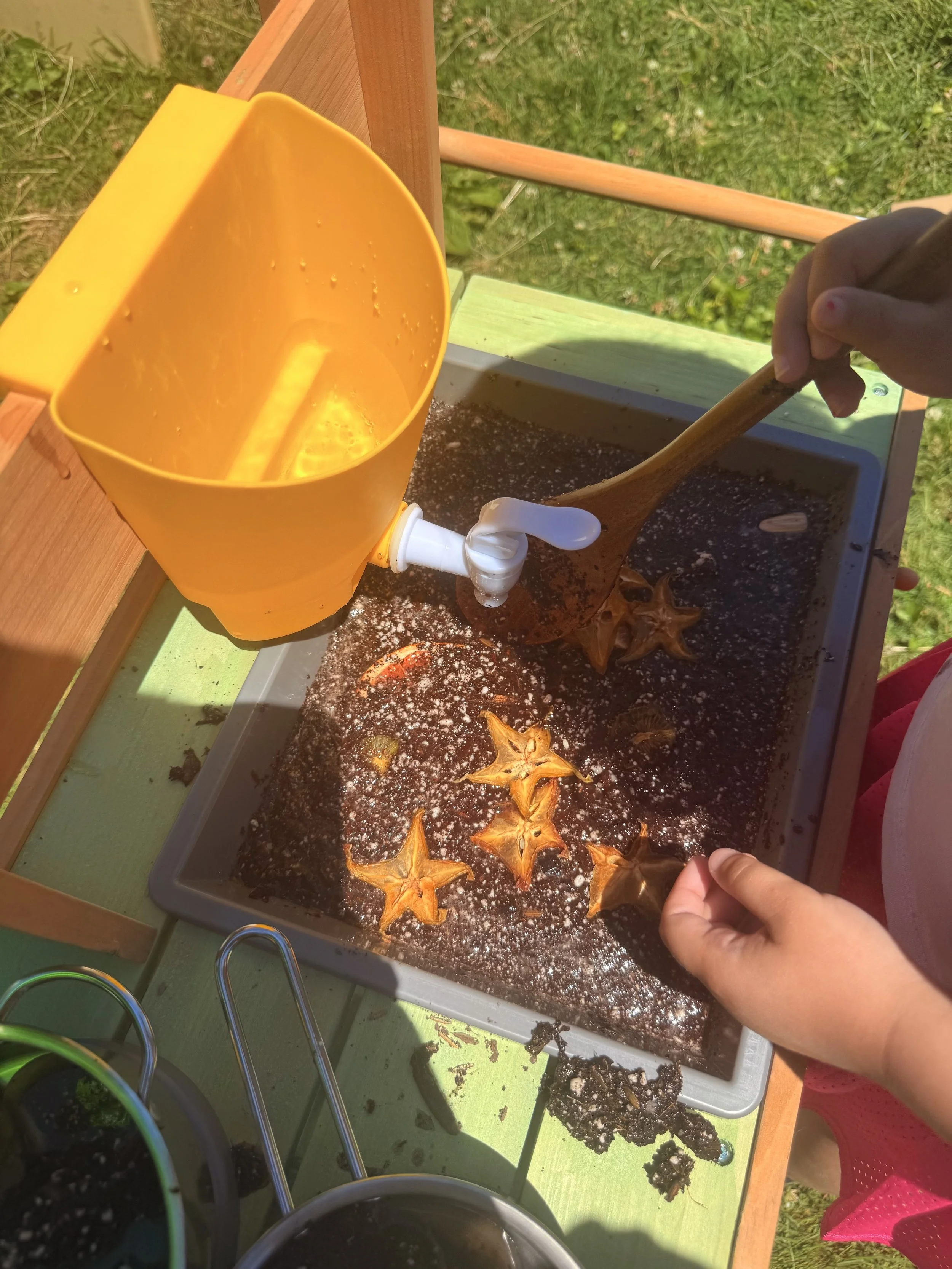 Child planting a seed in the soil using a small gardening trowel, with gardening supplies on a table outdoors.