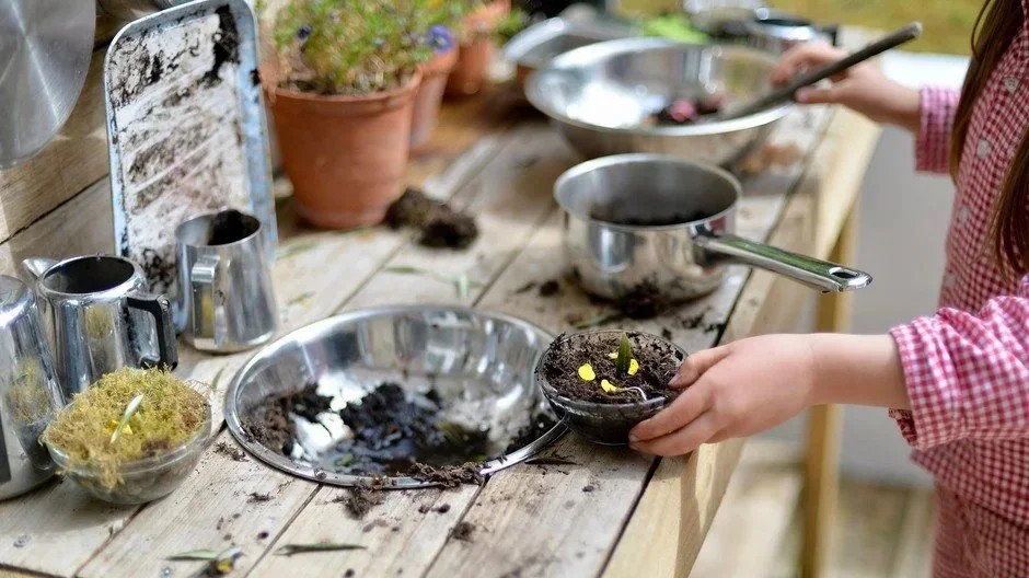 A person in a red checkered shirt is planting yellow flowers in a small bowl of soil, on a wooden table with gardening tools, pots, and pots with plants nearby.