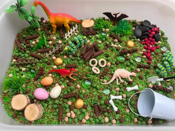 A miniature dinosaur scene with toy dinosaurs, plastic trees, colorful rocks, and fake plants, set on a bed of green sprinkles and chocolate pieces, resembling a prehistoric landscape.
