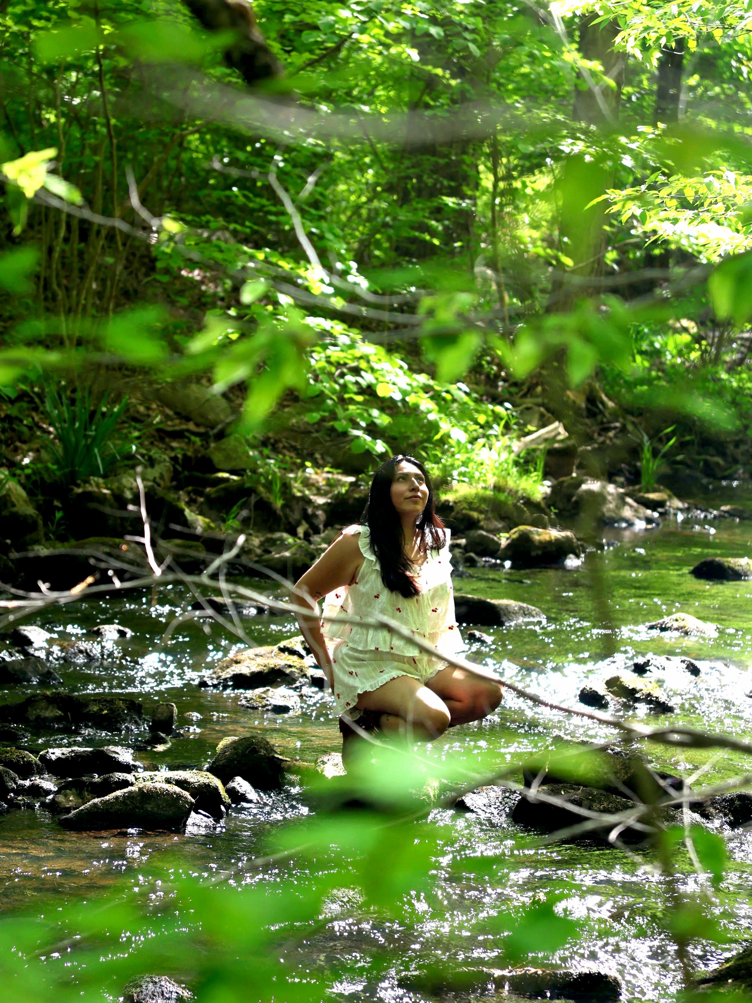 A woman kneeling in a shallow creek surrounded by lush green trees and foliage, with sunlight filtering through the leaves.