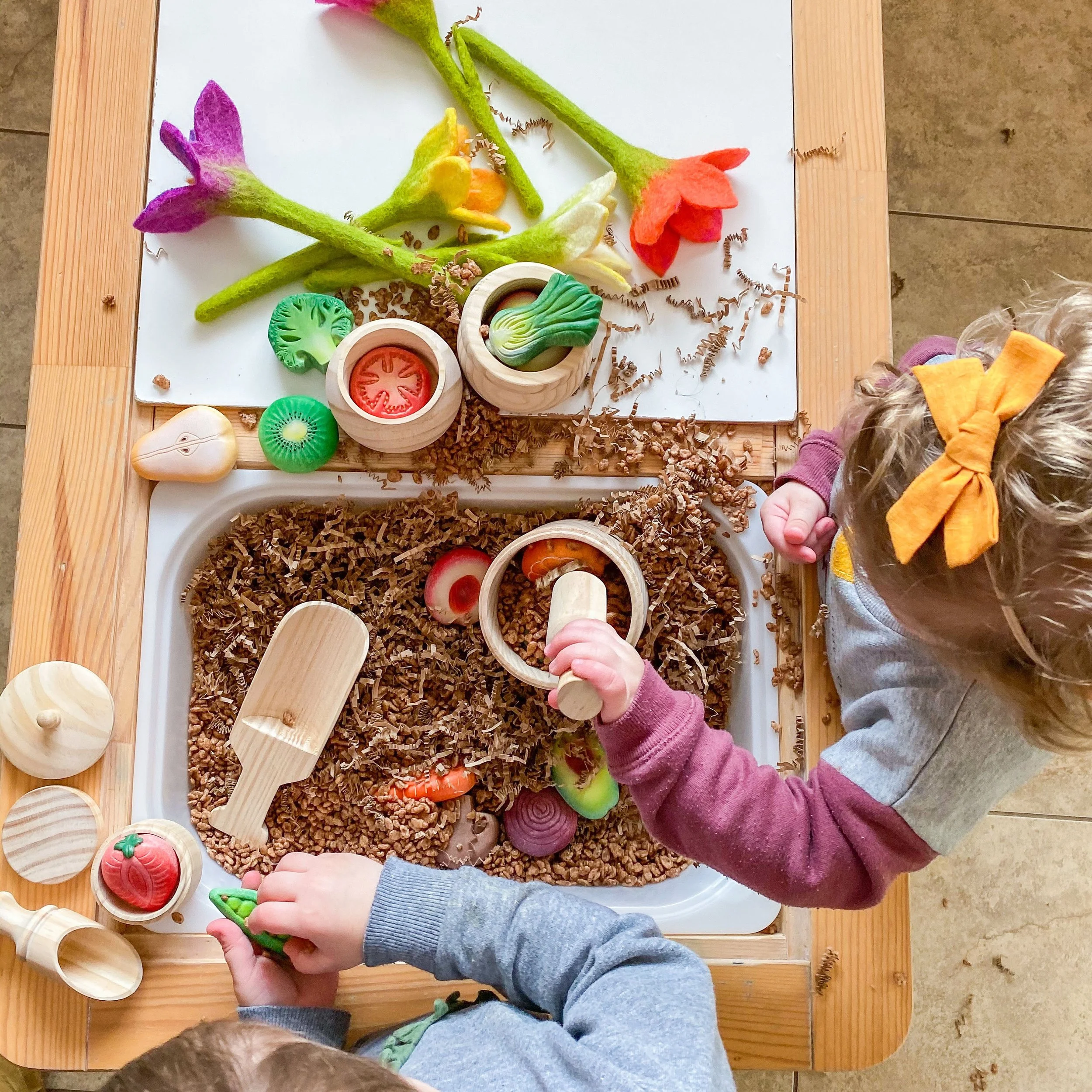 Children playing a tabletop game with felt and wooden food and kitchenware on a white surface.