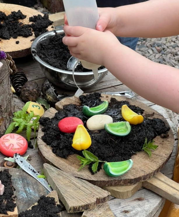 Child's hands pouring liquid into a plant pot filled with soil and colorful candy-like stones on a wooden surface.