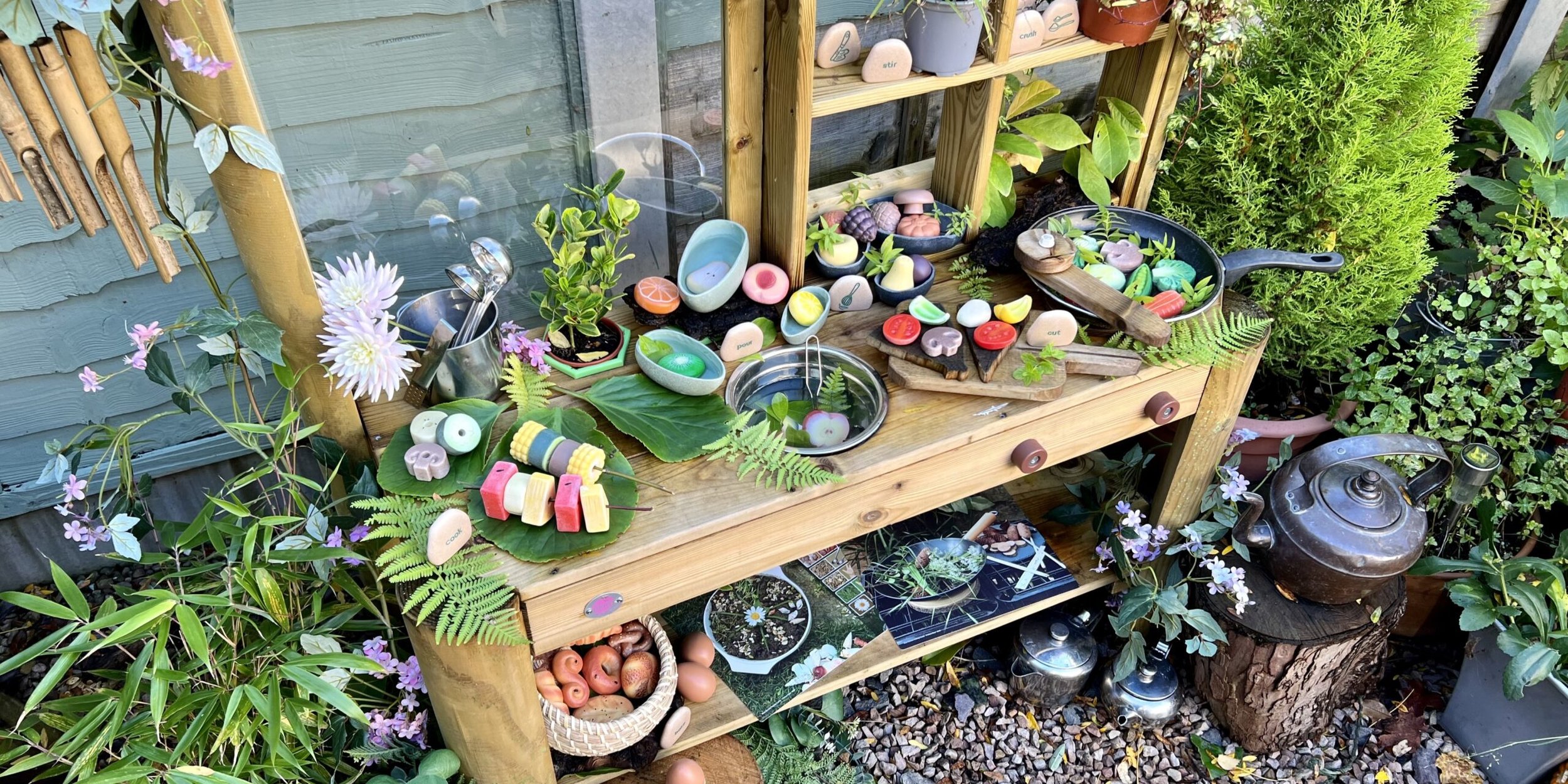 A wooden garden table decorated with colorful, playful stones resembling food and other items, surrounded by plants and flowers, with a teapot and various gardening tools.