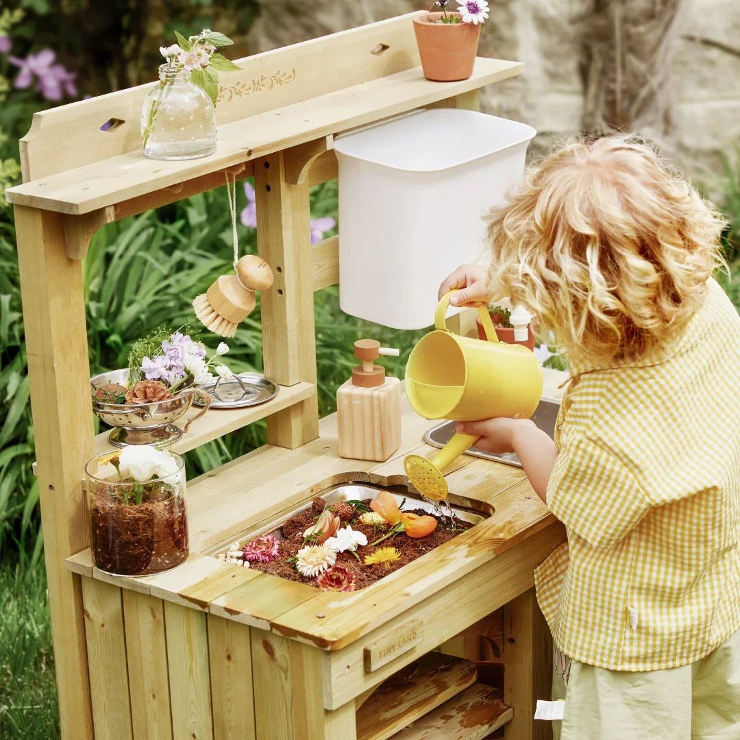 Child with curly hair playing at a miniature outdoor kitchen set, pouring water from a yellow watering can into a flower bed, surrounded by plants and garden decorations.
