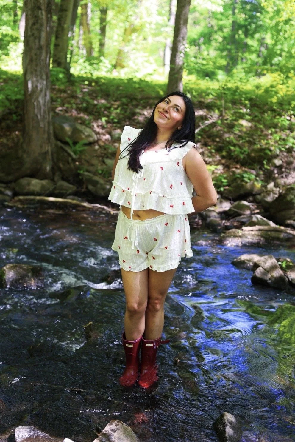A woman standing in a stream in a forest, smiling, wearing a cream-colored pajama set with cherry prints and red rain boots.