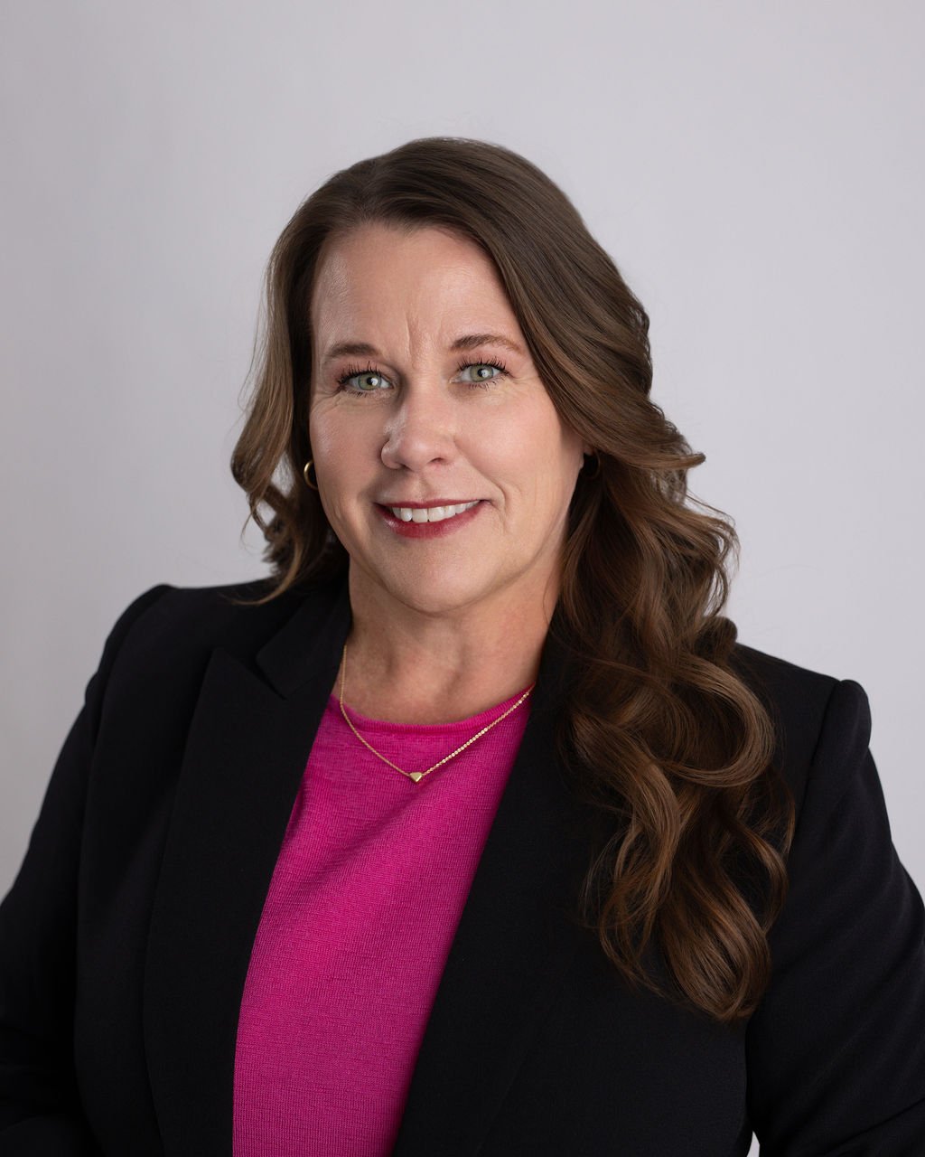 Professional woman with long brown hair, wearing a black blazer and pink top, smiling against a light grey background.