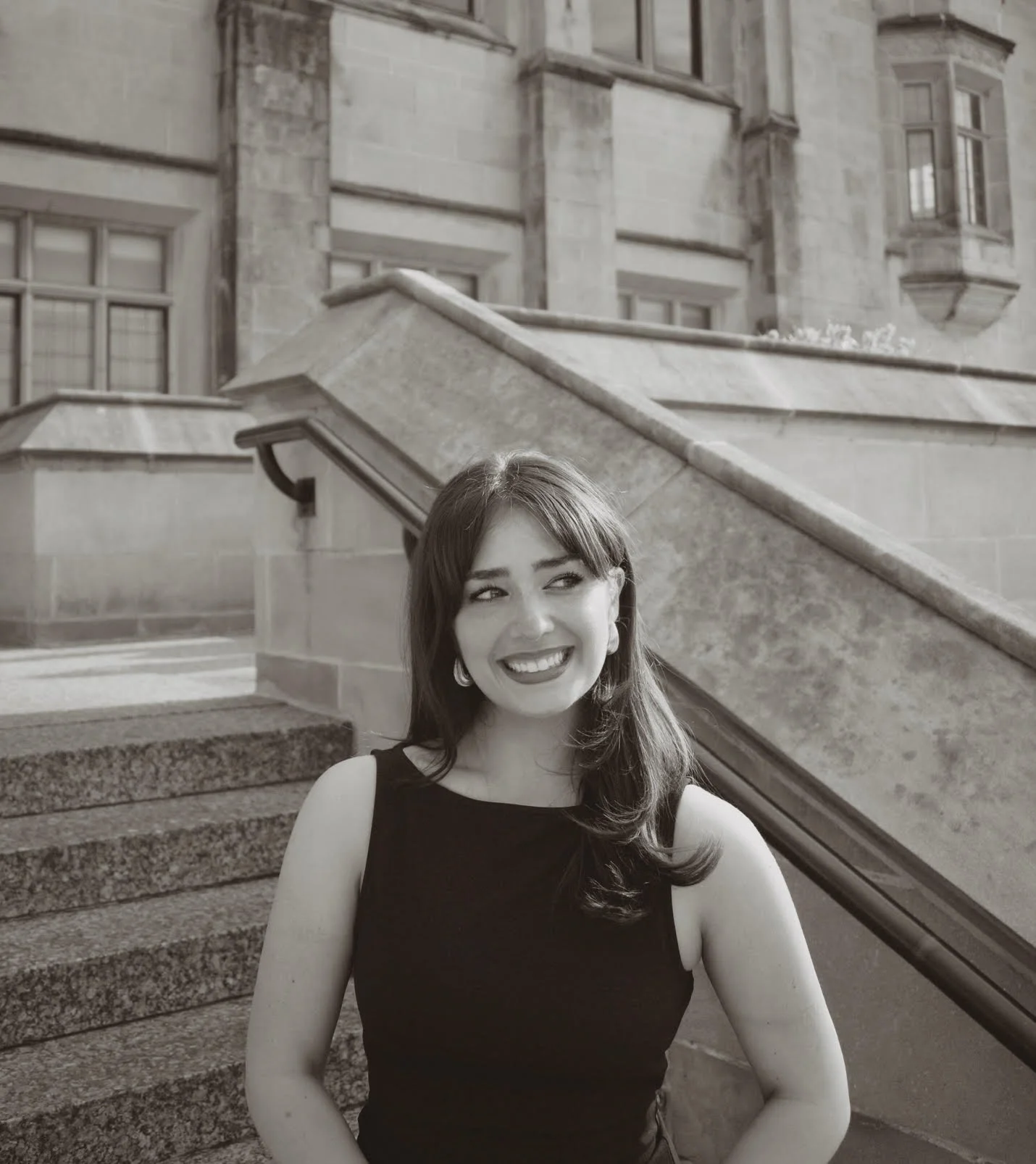 A young woman with shoulder-length hair and a big smile, wearing a sleeveless black top, standing outdoors in front of stone stairs and a historic building with windows.