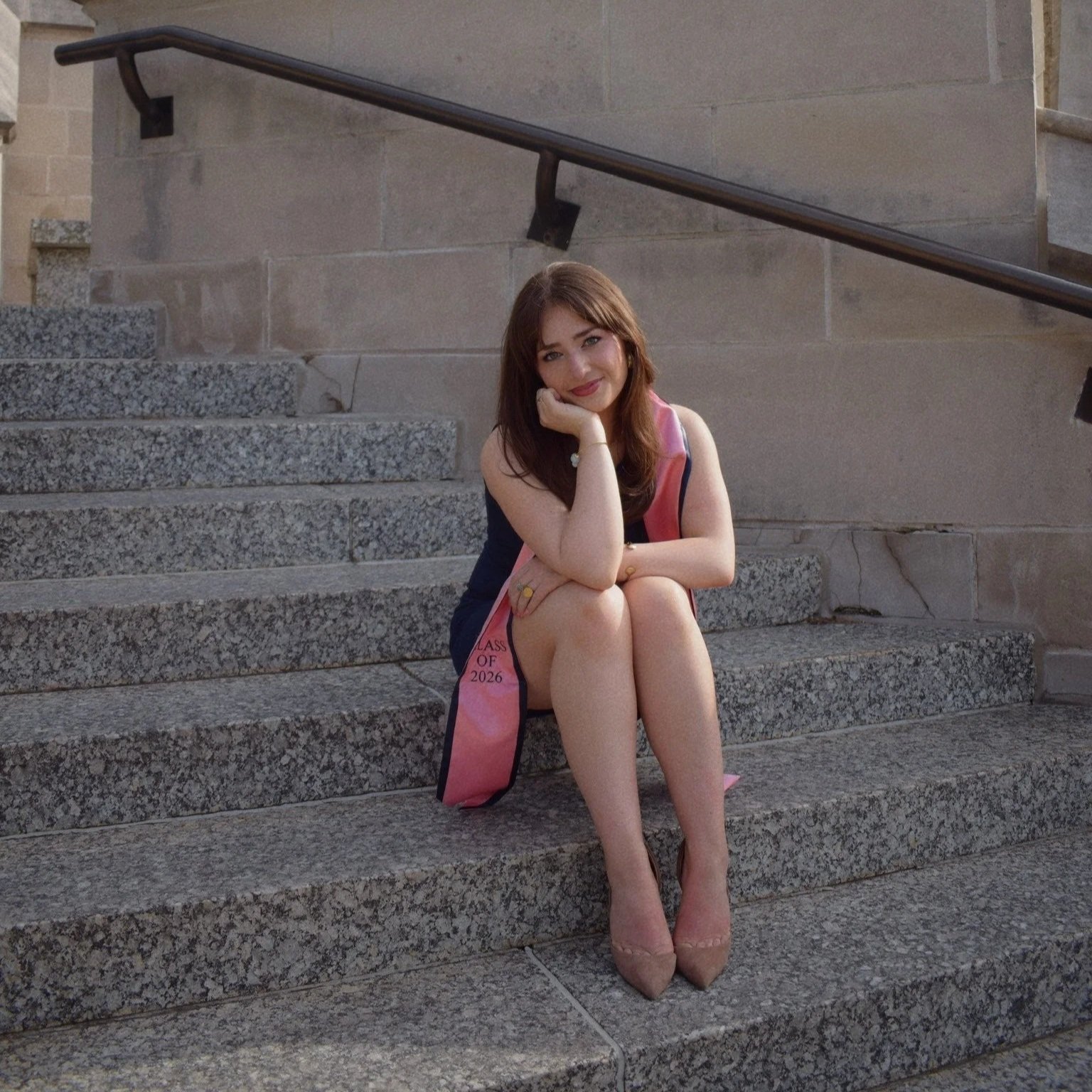 A young woman in a black dress and pink sash, sitting on gray stone steps, resting her chin on her hand, smiling at the camera, with a brick building wall behind her.
