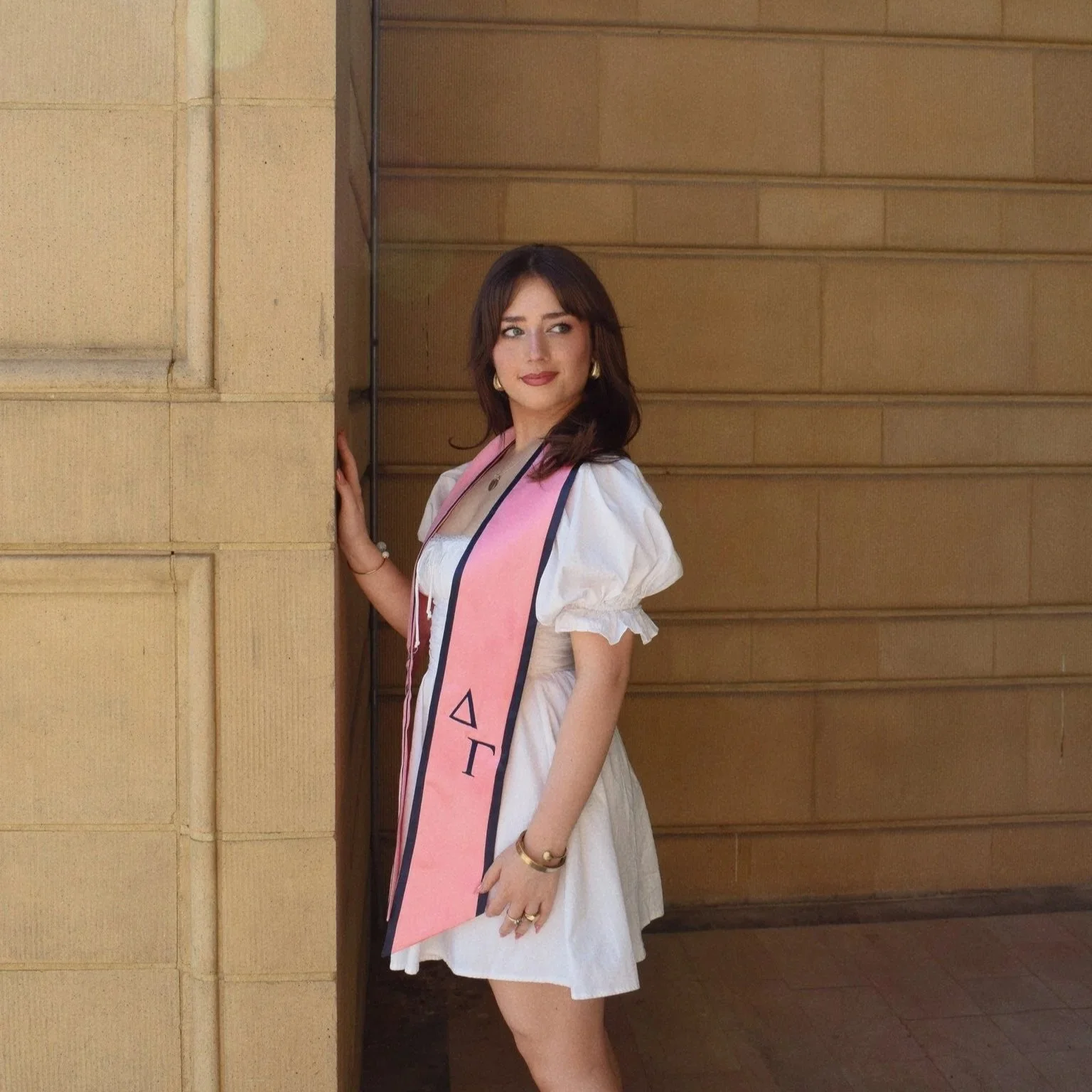 A young woman in a white dress with a pink sash stands beside a beige stone wall, looking to the side.