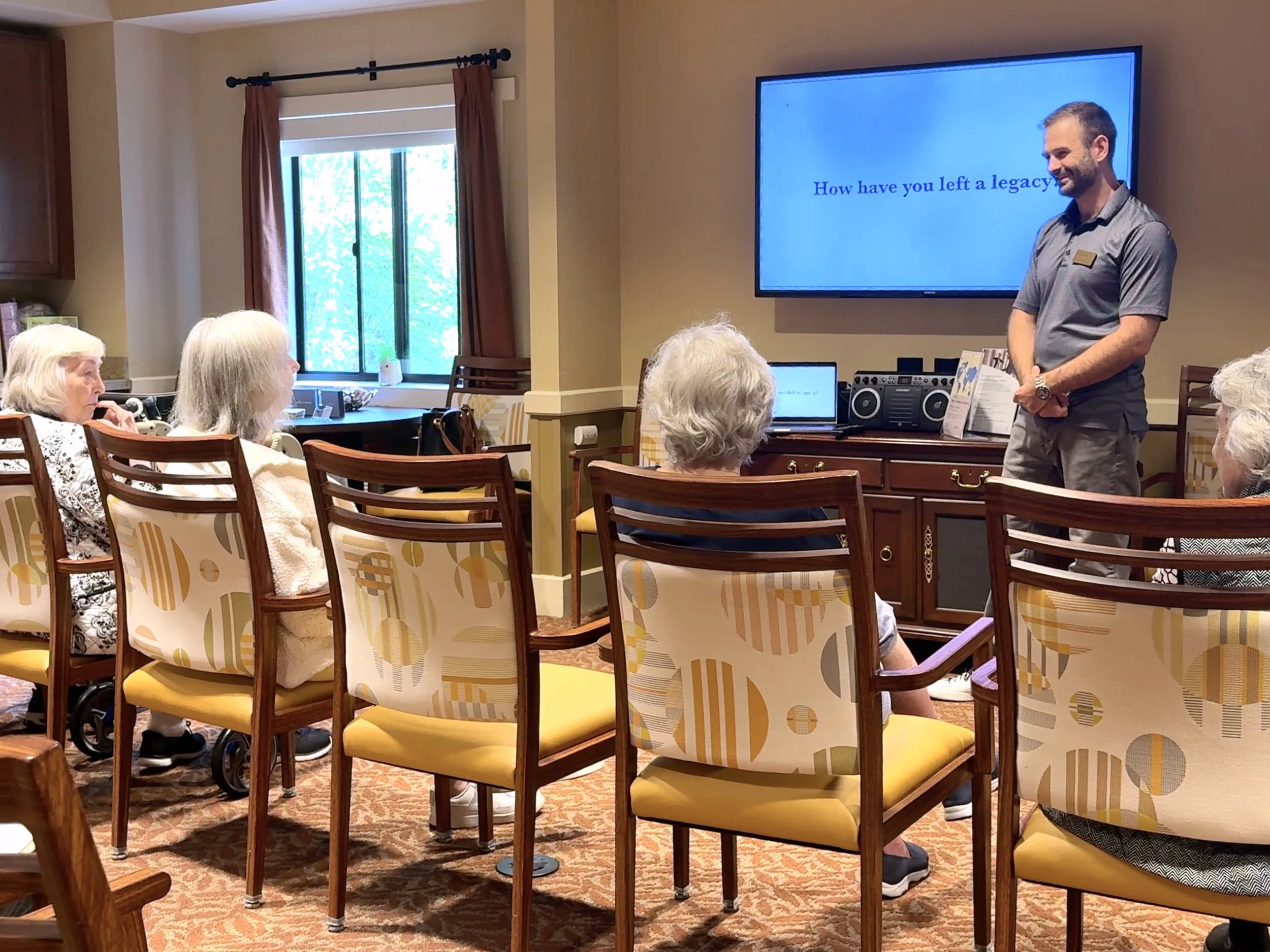 A group of seniors attending a presentation in a cozy room with yellow walls, facing a man standing next to a large screen that reads 'How have you left a legacy?'. The room has a window with curtains, and there are chairs with patterned cushions.