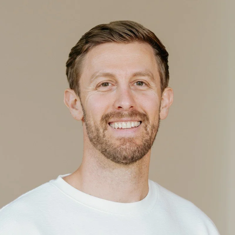A smiling man with short brown hair, a beard, and wearing a white shirt, standing against a plain background.