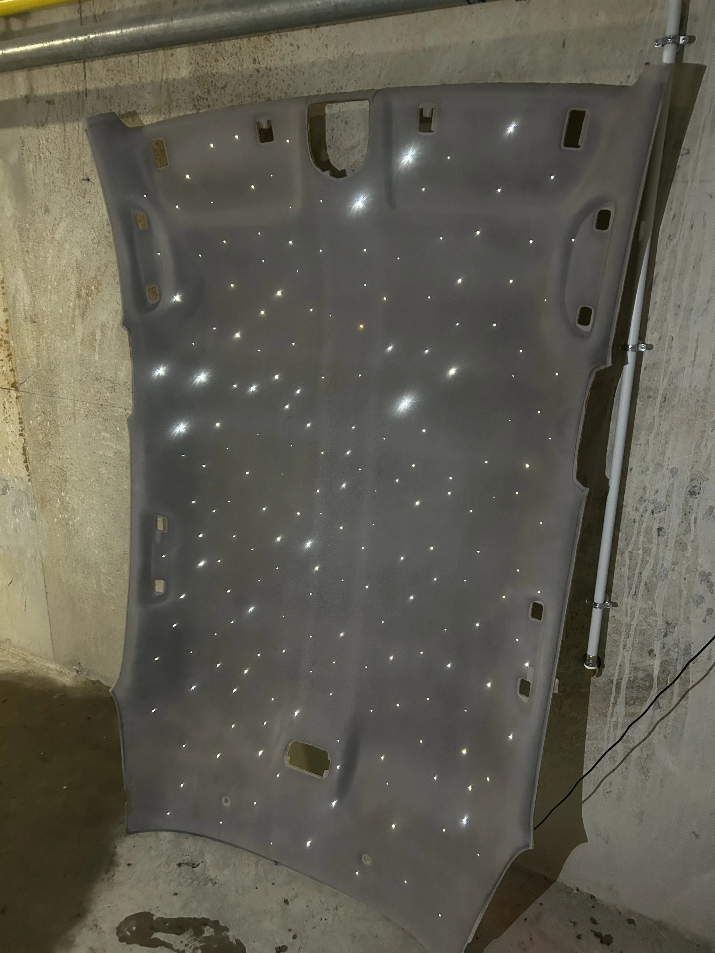 Une grande pièce métallique, peut-être une pièce de carrosserie, avec de nombreux petits trous lumineux semblables à des étoiles. Elle est posée à même le sol dans un espace industriel ou en construction.