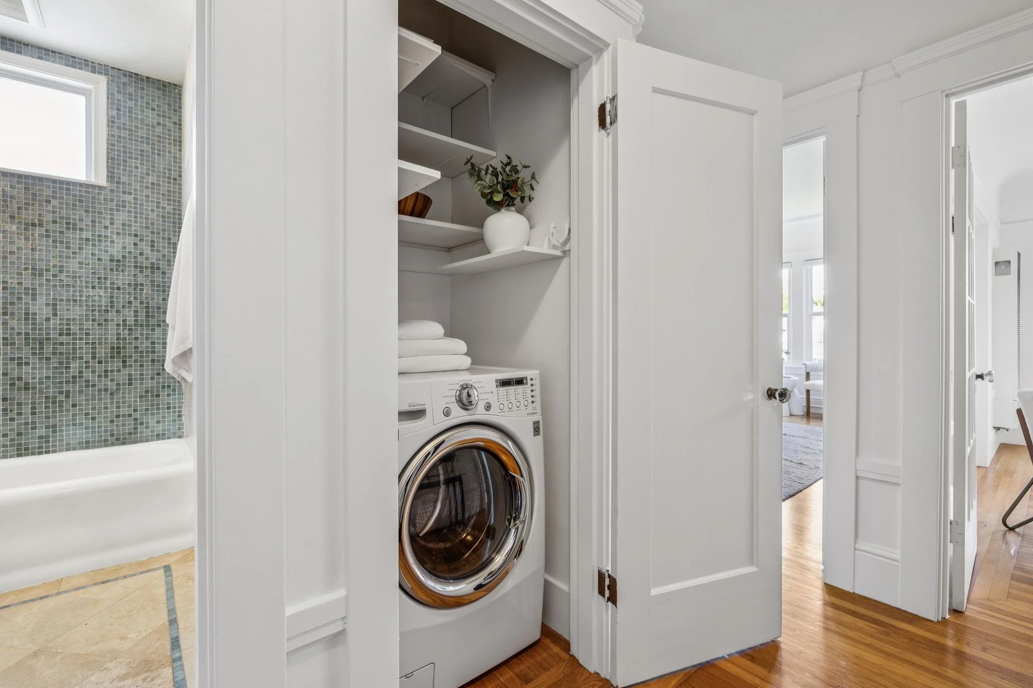 Laundry area with a stacked washer and dryer behind a white cabinet door, adjacent to a bathroom with a shower featuring small mosaic tiles.