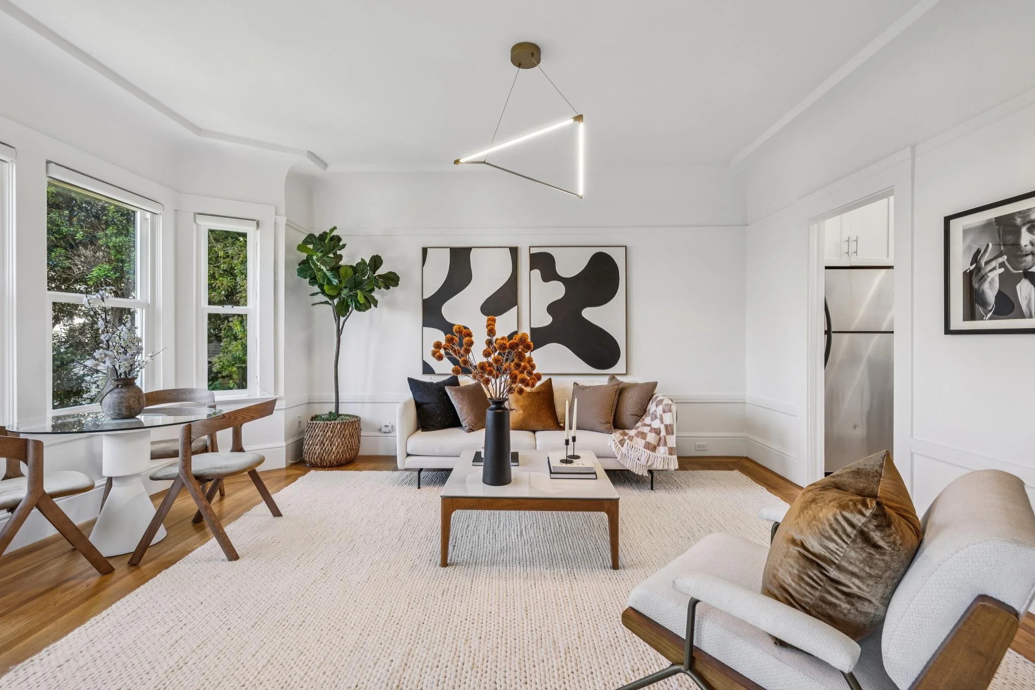 A modern living room with white walls and large windows, featuring a white sofa with brown and black cushions, abstract black and white artwork on the wall, a round wooden dining table with beige chairs, houseplants, a white coffee table with decorat