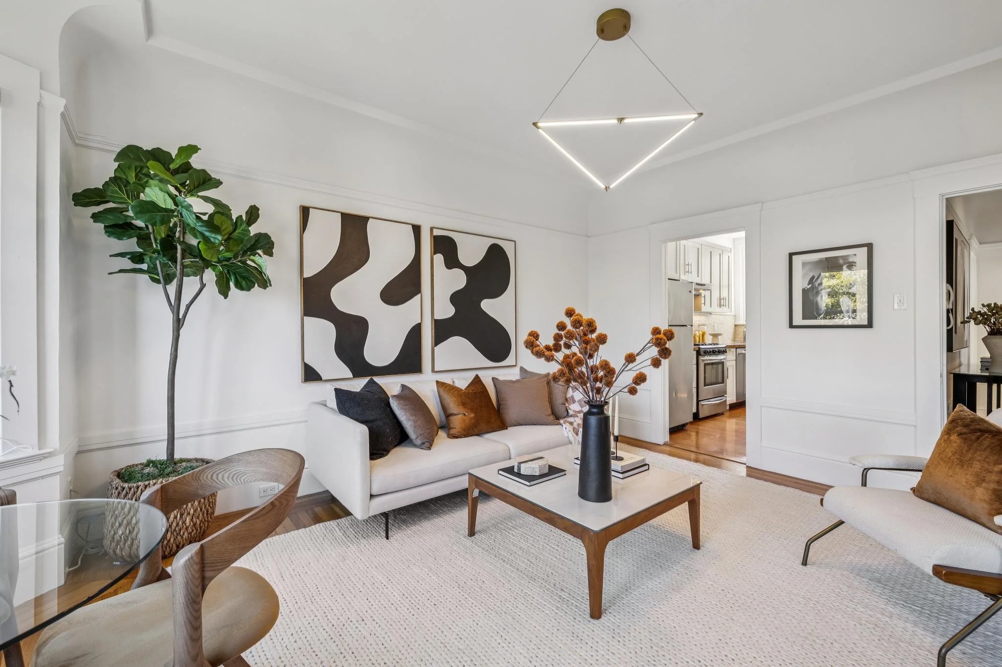 A living room with white walls and ceiling, decorated with modern black and white abstract art, a white sofa with brown and black pillows, a black vase with dried orange flowers on a coffee table, a wooden side table, and a large green plant. There's