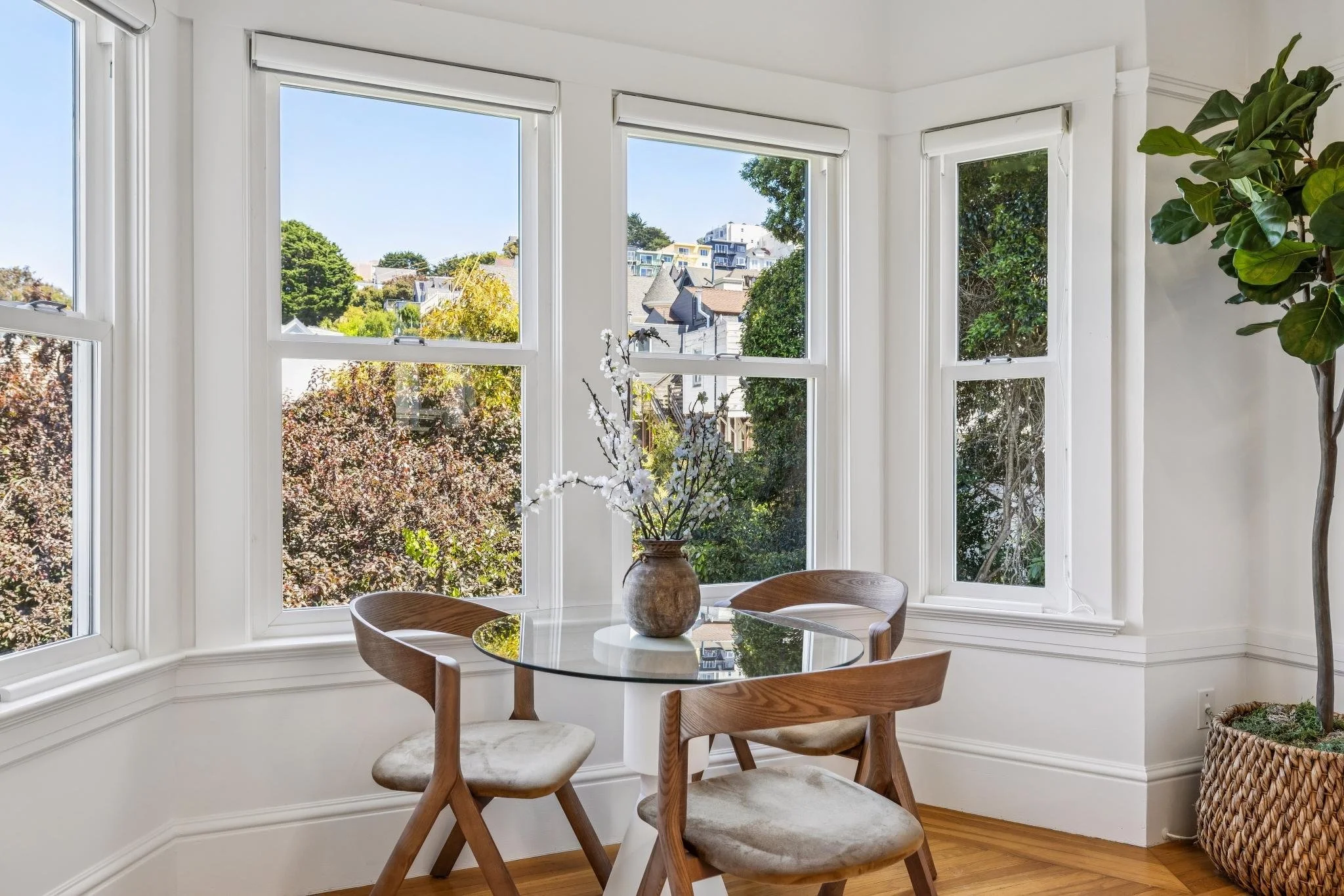 A bright kitchen corner with three large windows showing trees and neighboring buildings outside. There is a round glass-top table with a vase of white flowers and three wooden chairs with upholstered seats. A tall potted plant is in the corner.