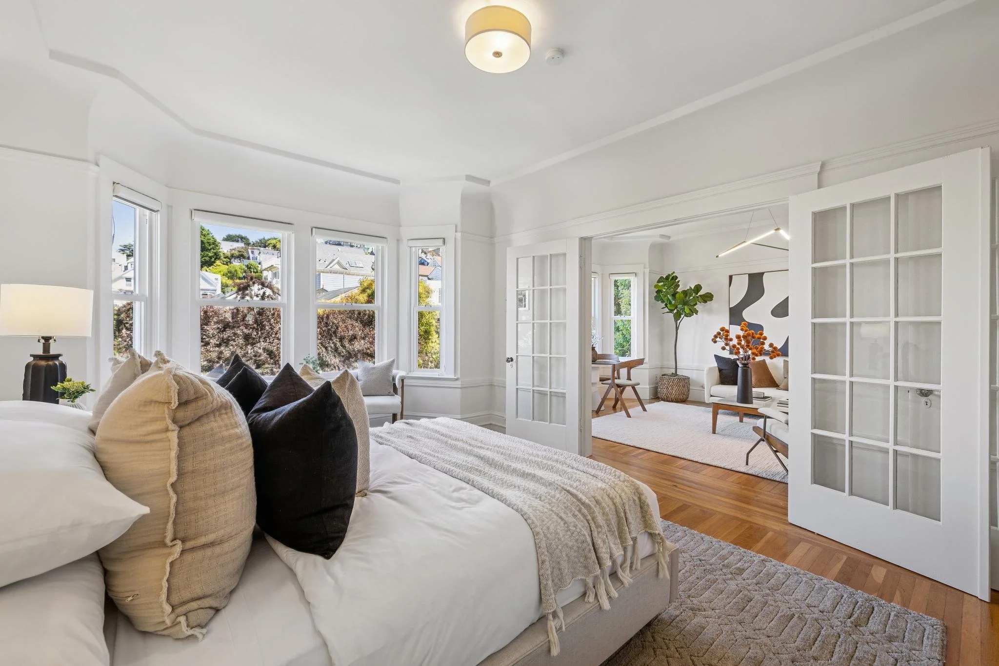 Bright living room with white walls, large windows, a cozy couch with assorted cushions, and a view of neighboring houses and trees outside.