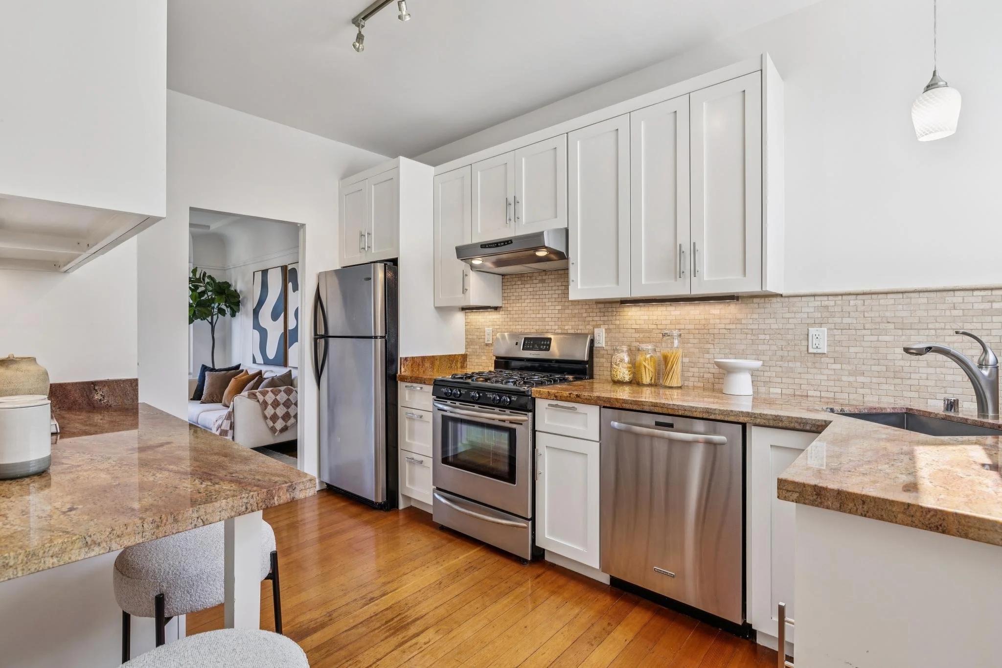 A modern kitchen with white cabinets, stainless steel appliances, granite countertops, and a beige brick backsplash. There is a small living area visible through an open doorway.