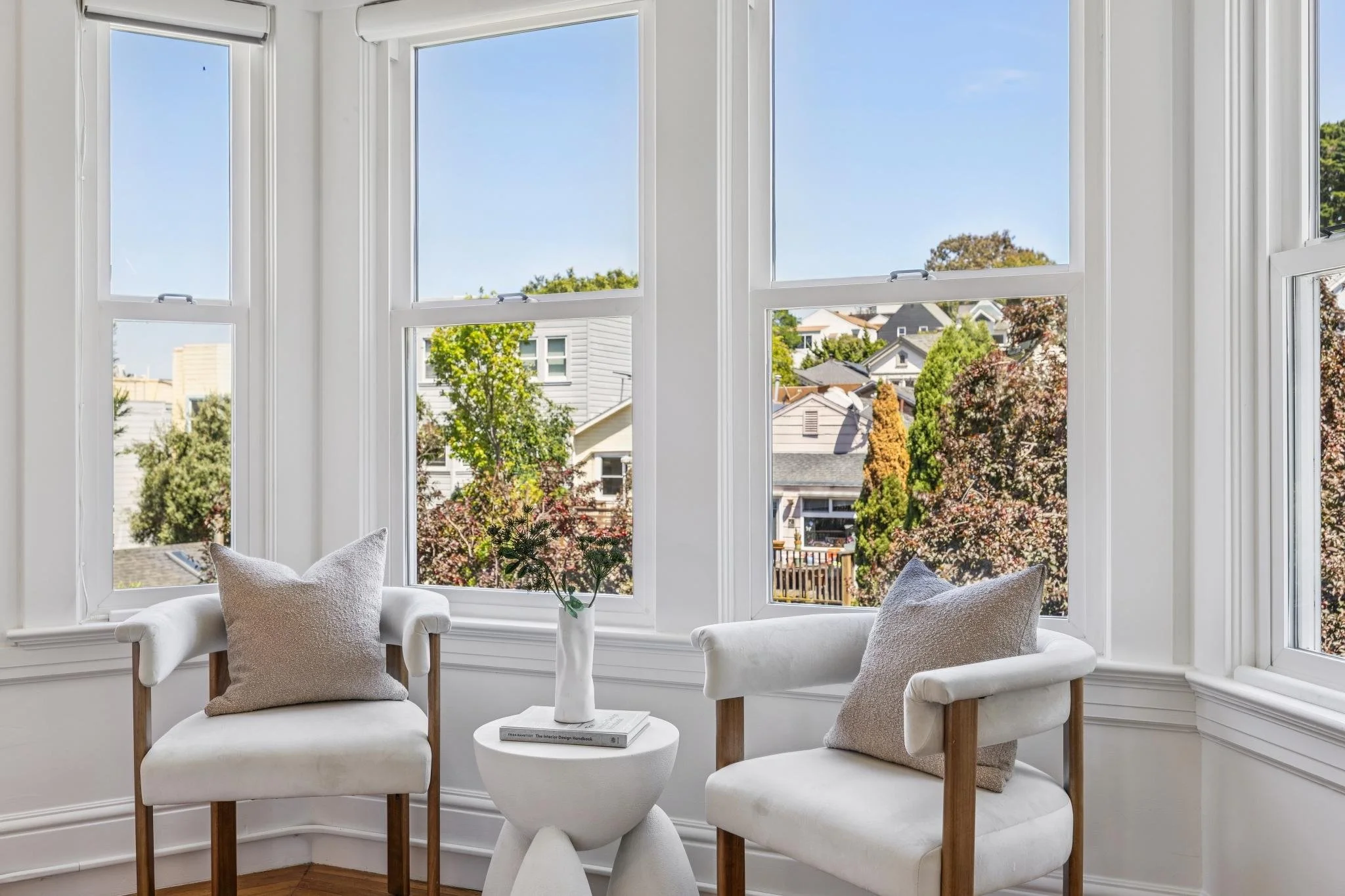 A cozy interior scene featuring three white chairs with beige cushions around a small white round table with a vase of flowers and a book, set in front of a large bay window with a view of trees and houses in a suburban neighborhood.