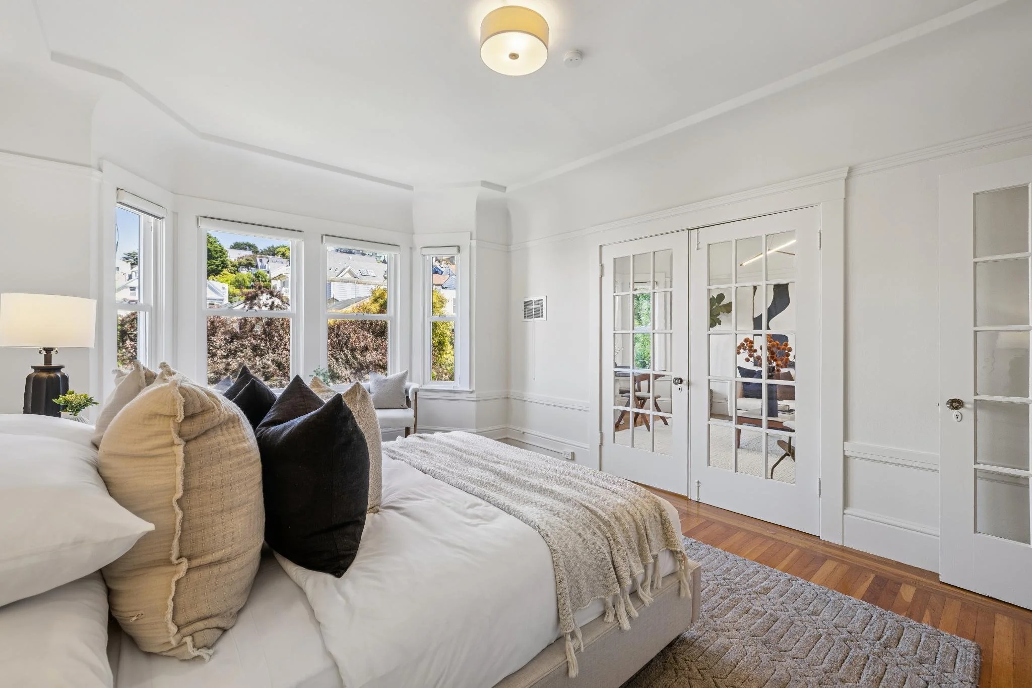 Bright living room with a white sofa, multiple pillows, large windows with views of houses and trees, and a set of French doors leading to another room.