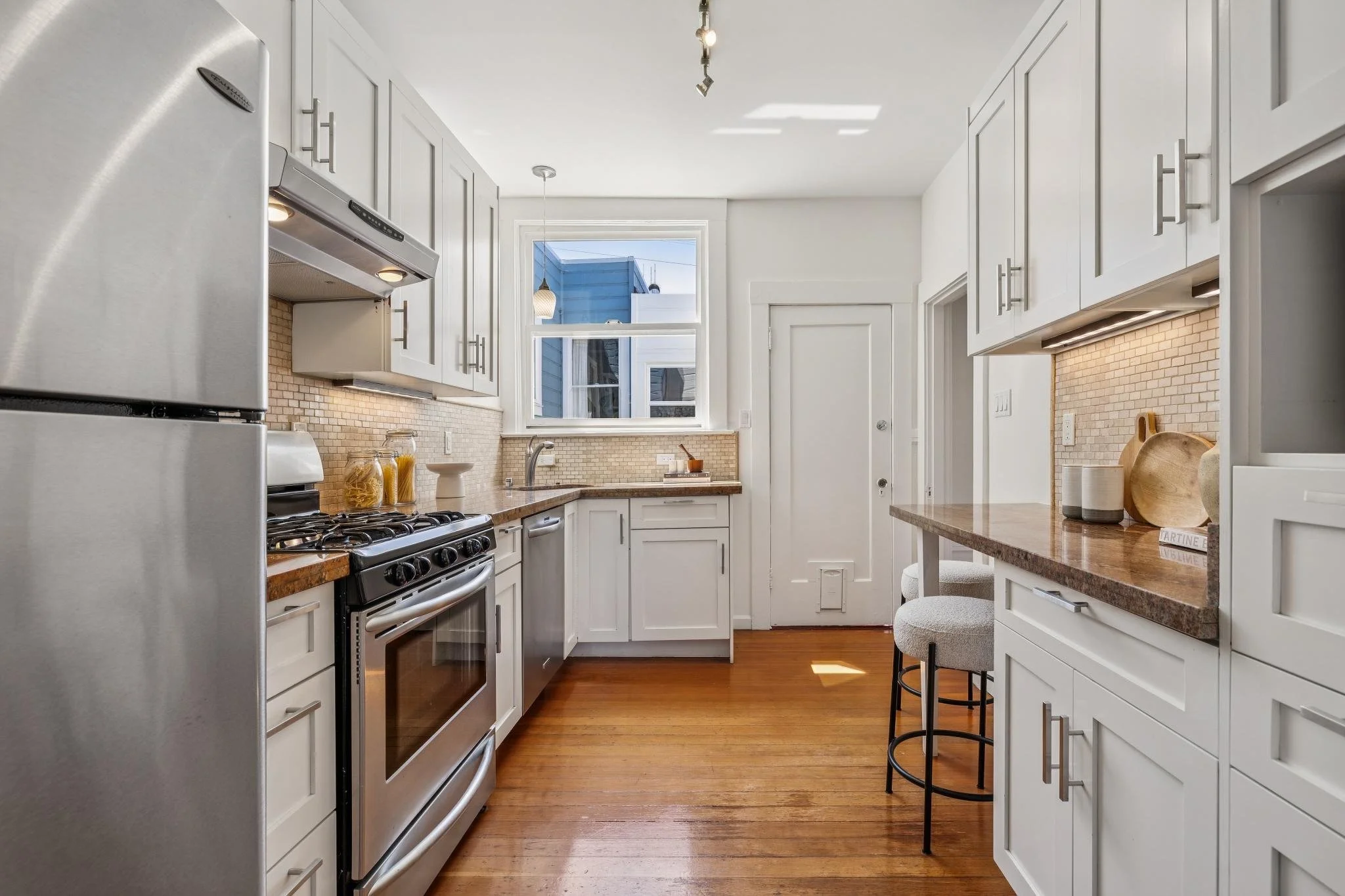 Bright, modern kitchen with white cabinets, a wooden countertop, stainless steel appliances, and a window overlooking neighboring buildings.