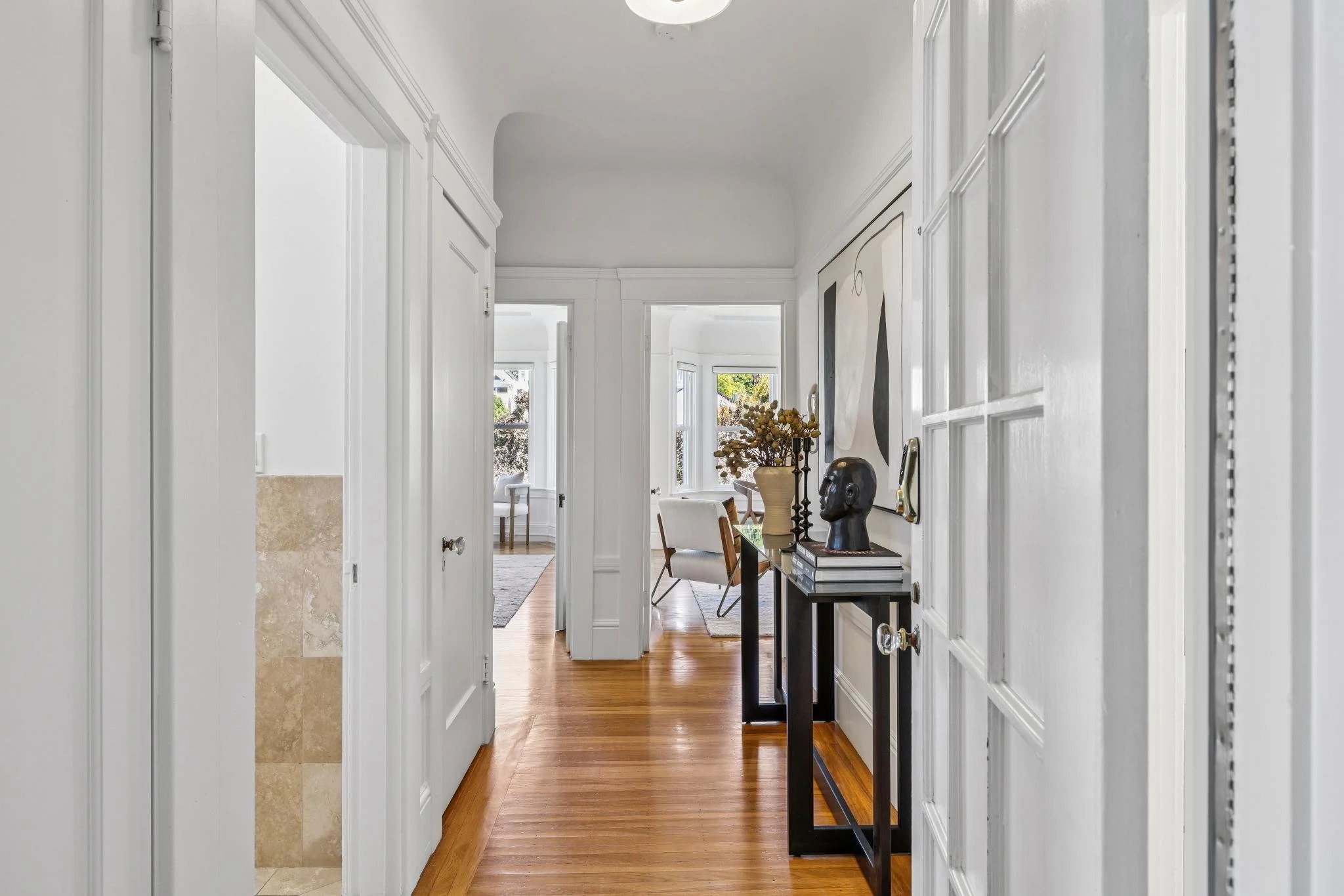 Hallway leading into a bright living room with large windows, artwork, a table with decorative items, and chairs.