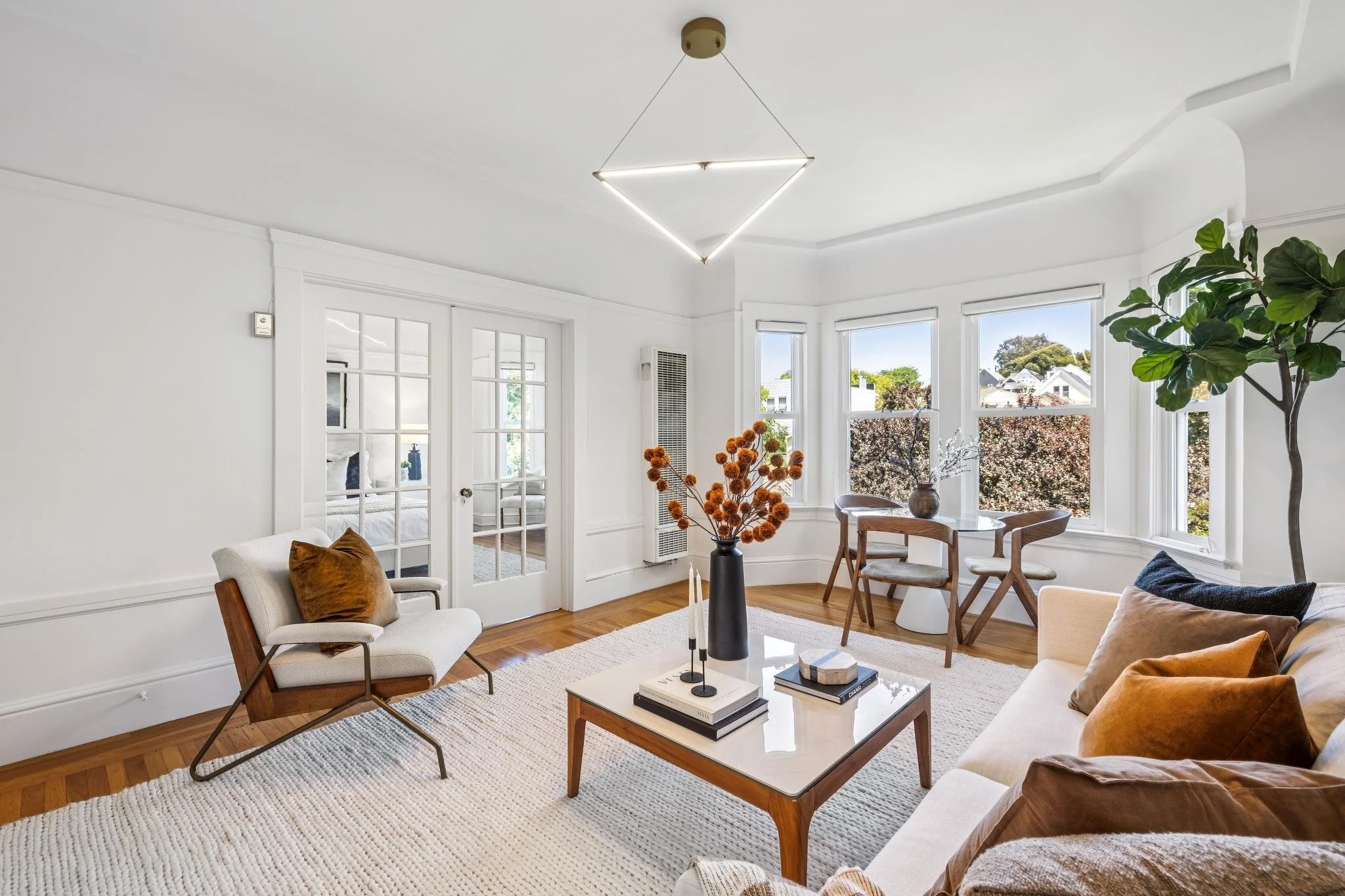 Bright living room with large windows, white walls, wooden floor, and modern furniture including a beige sofa, white armchair with brown pillow, wooden chairs, and a glass coffee table with books and candles. Decor includes an orange flower arrangeme