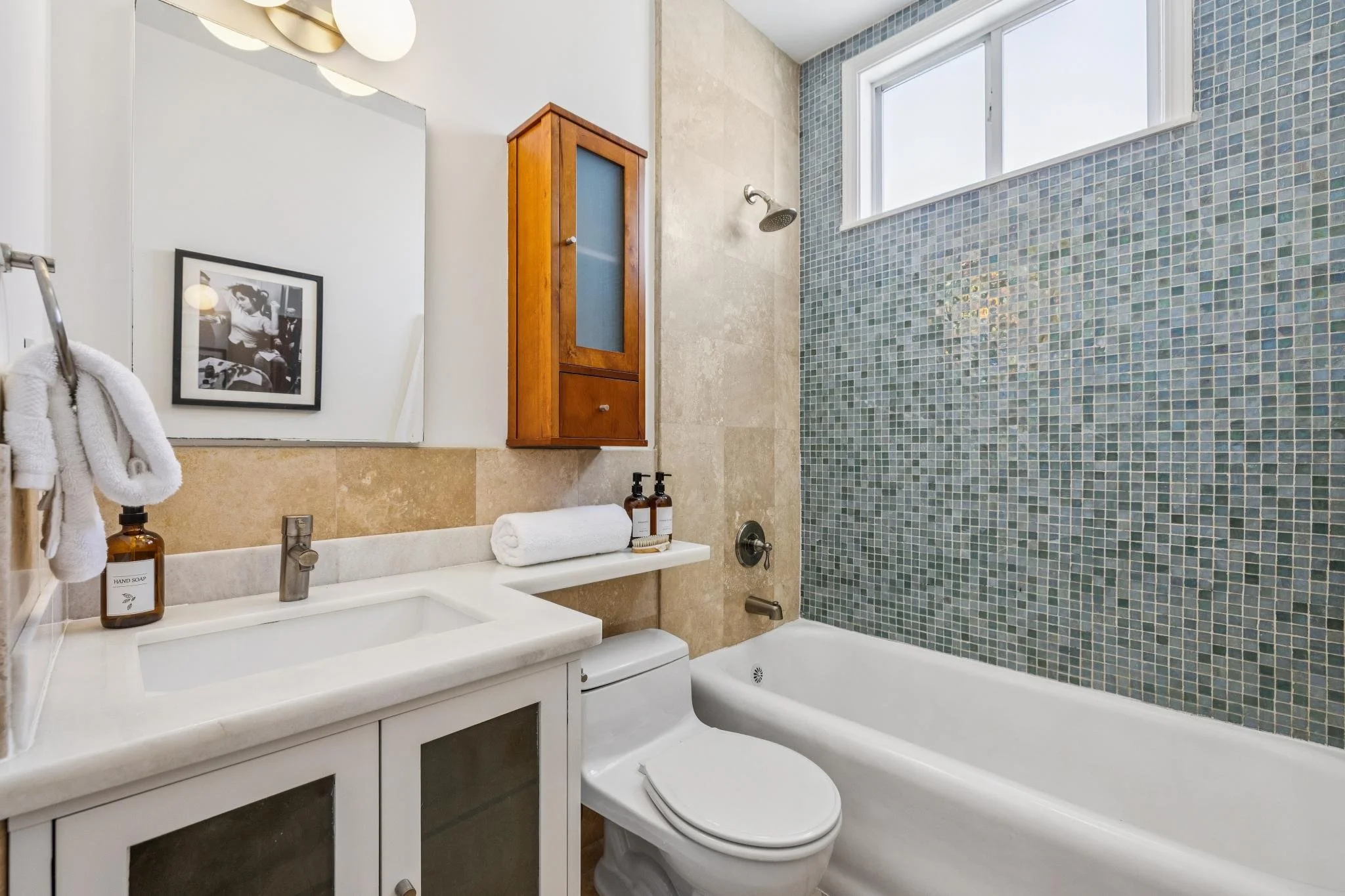 A clean bathroom with a white vanity, a large mirror, a framed black-and-white photo, wooden wall cabinet, towels, liquid soap bottles, a bathtub, and a tiled shower wall with a small window.