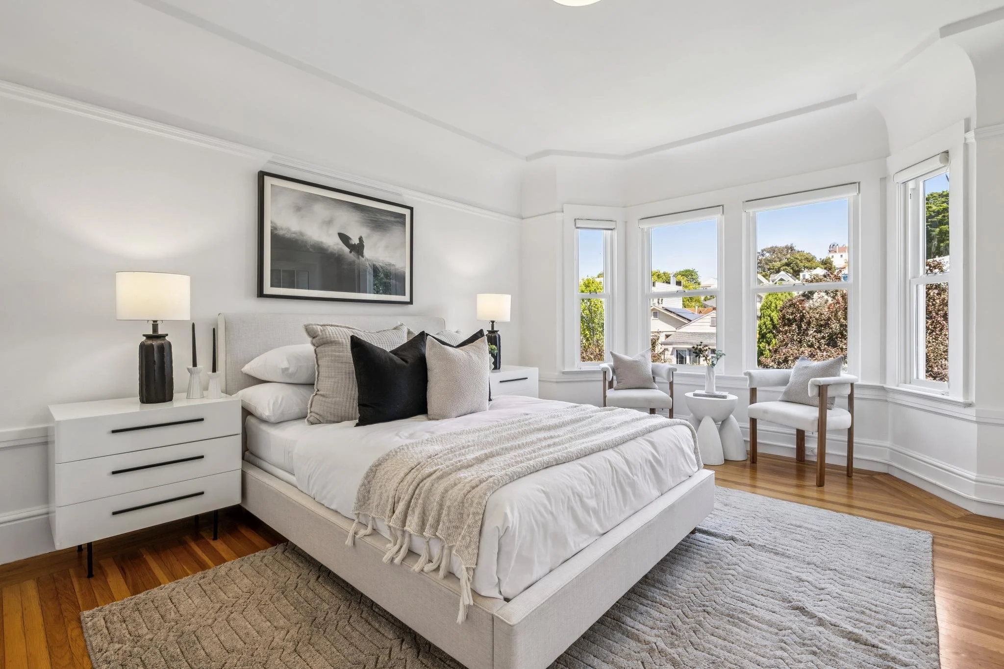 Bright bedroom with large bay windows, white walls, a white bed with neutral pillows, black and white abstract artwork, two white bedside tables with lamps, wooden flooring, and a beige rug.