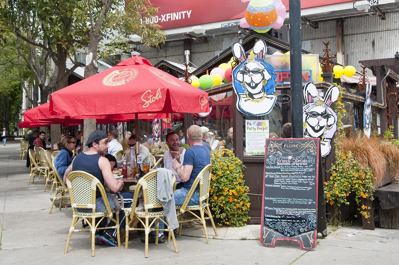People dining outdoors at a restaurant with red umbrellas decorated with the Stoli logo, a chalkboard menu, and bunny-themed decorations outside.