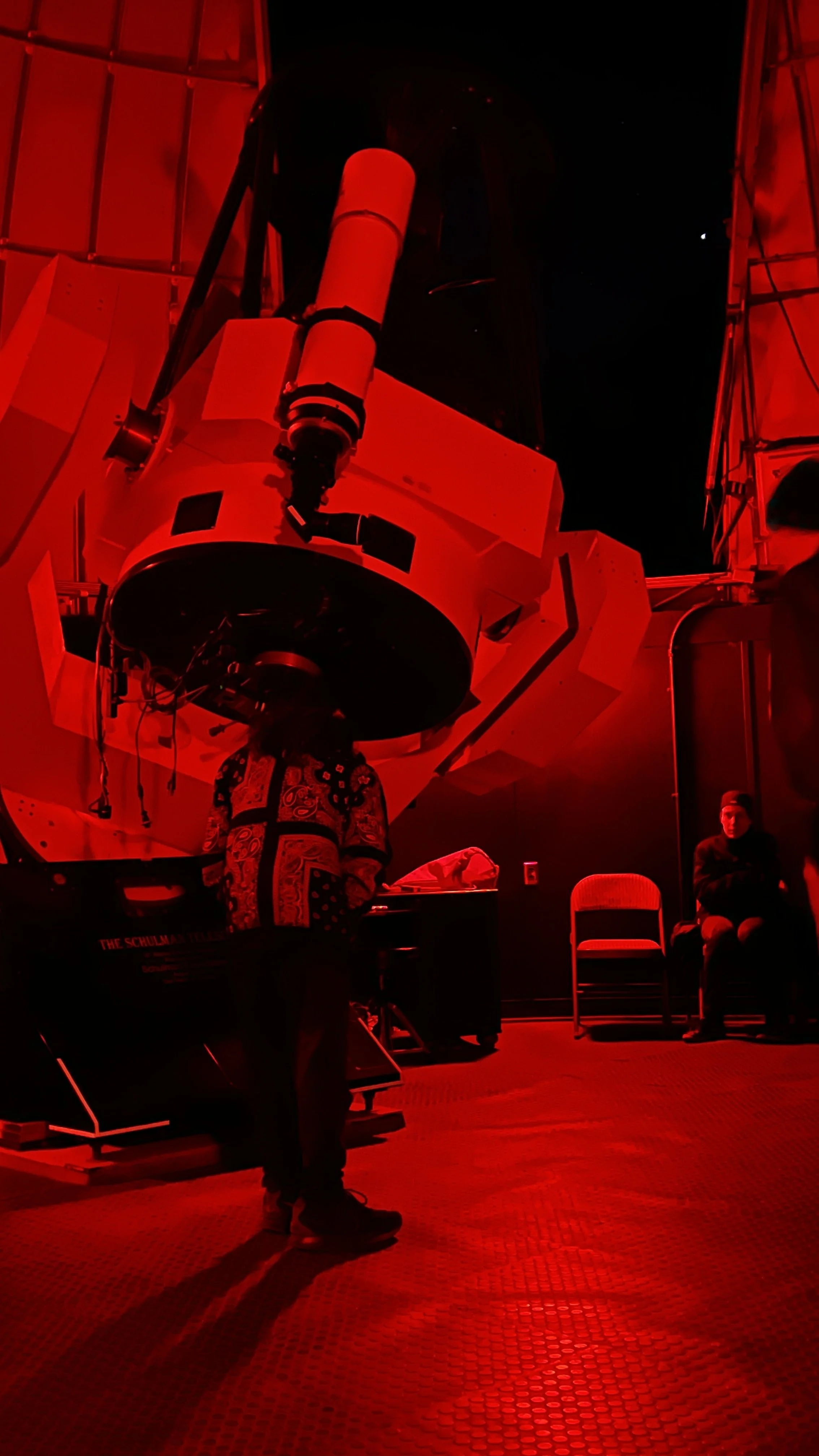 A large scientific telescope inside a building with red lighting, with a few people around it and chairs against the wall.
