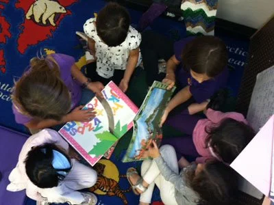A group of five children sitting on a colorful carpet, looking at picture books together.