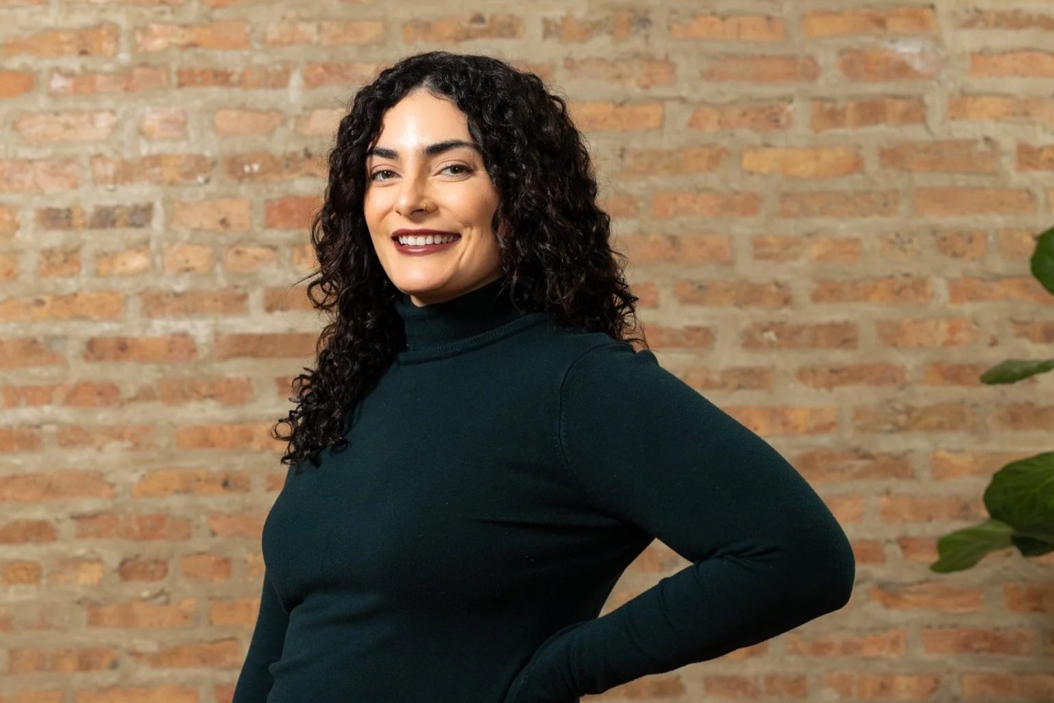Amanda standing in front of a brick wall wearing a dark green turtle neck, she's smiling, and wearing her hair down and curly.