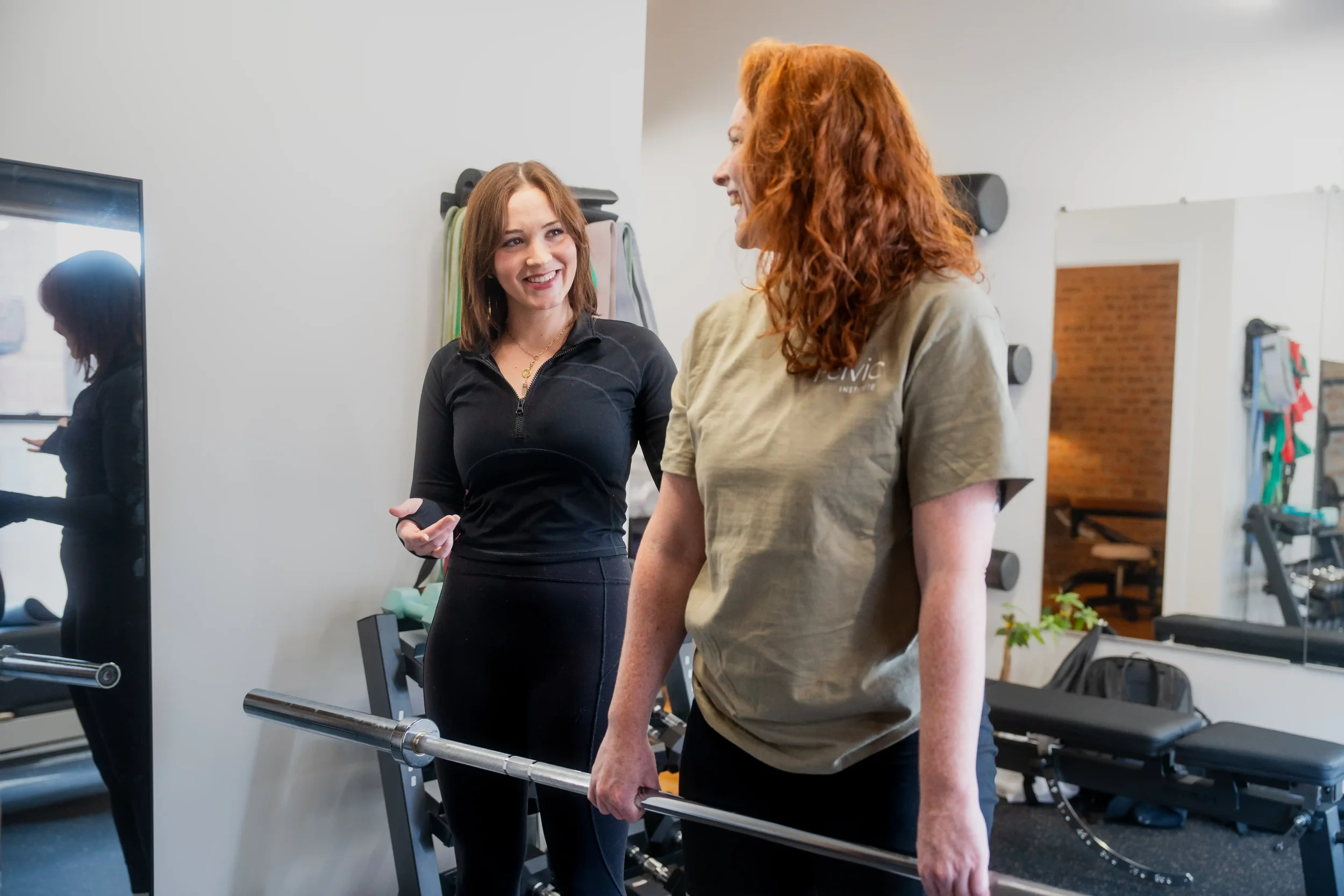 A person with red hair holding a barbell working with their physical therapist. Her physical therapist is smiling and supportive as she coaches the woman through a deadlift.