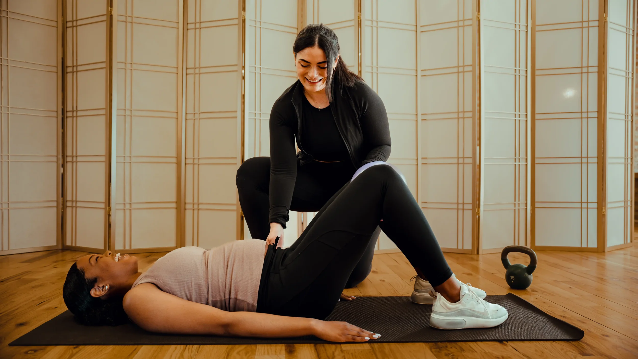 A woman is assisting another woman during a fitness session in a room with wooden floors and decorative folding screens. The woman on the floor is lying on a yoga mat, smiling, with her knees bent and feet flat on the ground. The trainer is standing over her, holding her ankles and smiling, as they perform a fitness exercise together. There is a kettlebell on the floor nearby.