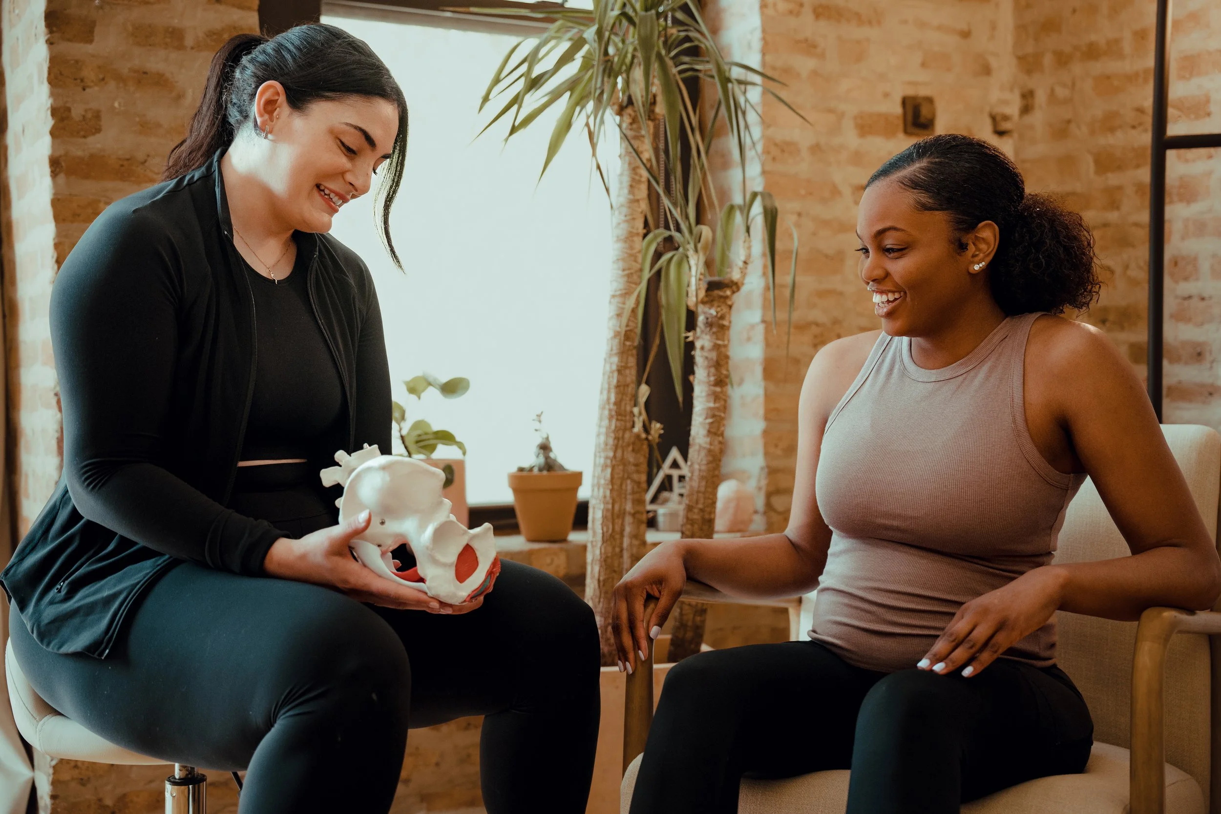 A physical therapists holds a model of a pelvis, while her patient sits comfortably in the chair next to her. In the background we see plants lining a brick wall.