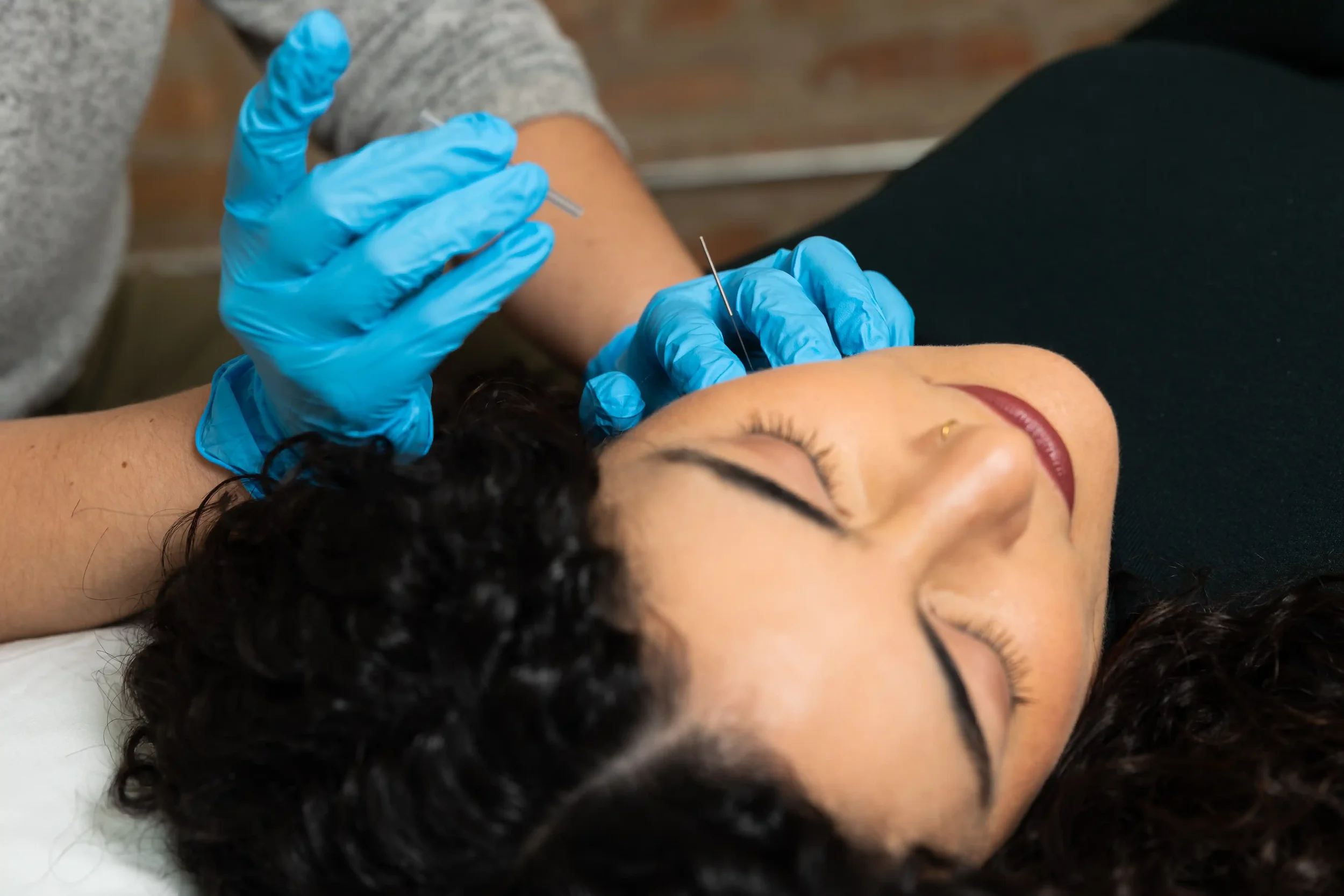 A woman receiving an injection in her upper arm, lying down with eyes closed, wearing a black top, with someone in blue gloves administering the shot.