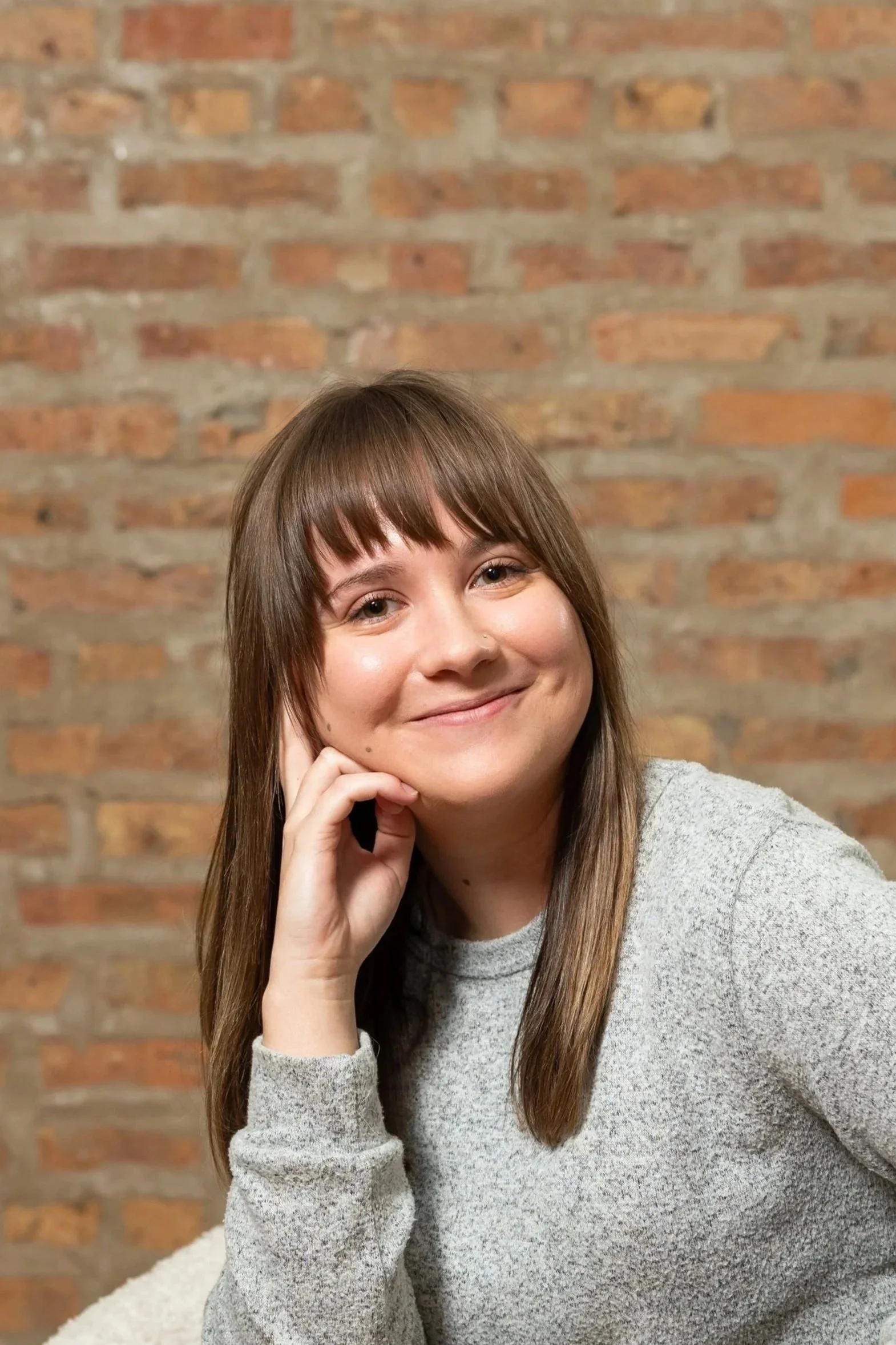 A young woman with long straight hair wearing a gray T-shirt, smiling, against a blurred brick wall background.