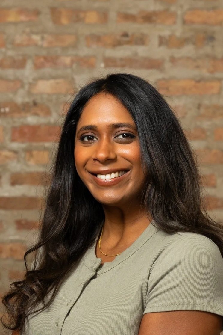 A woman with long black hair and a tan shirt smiling in front of a brick wall.