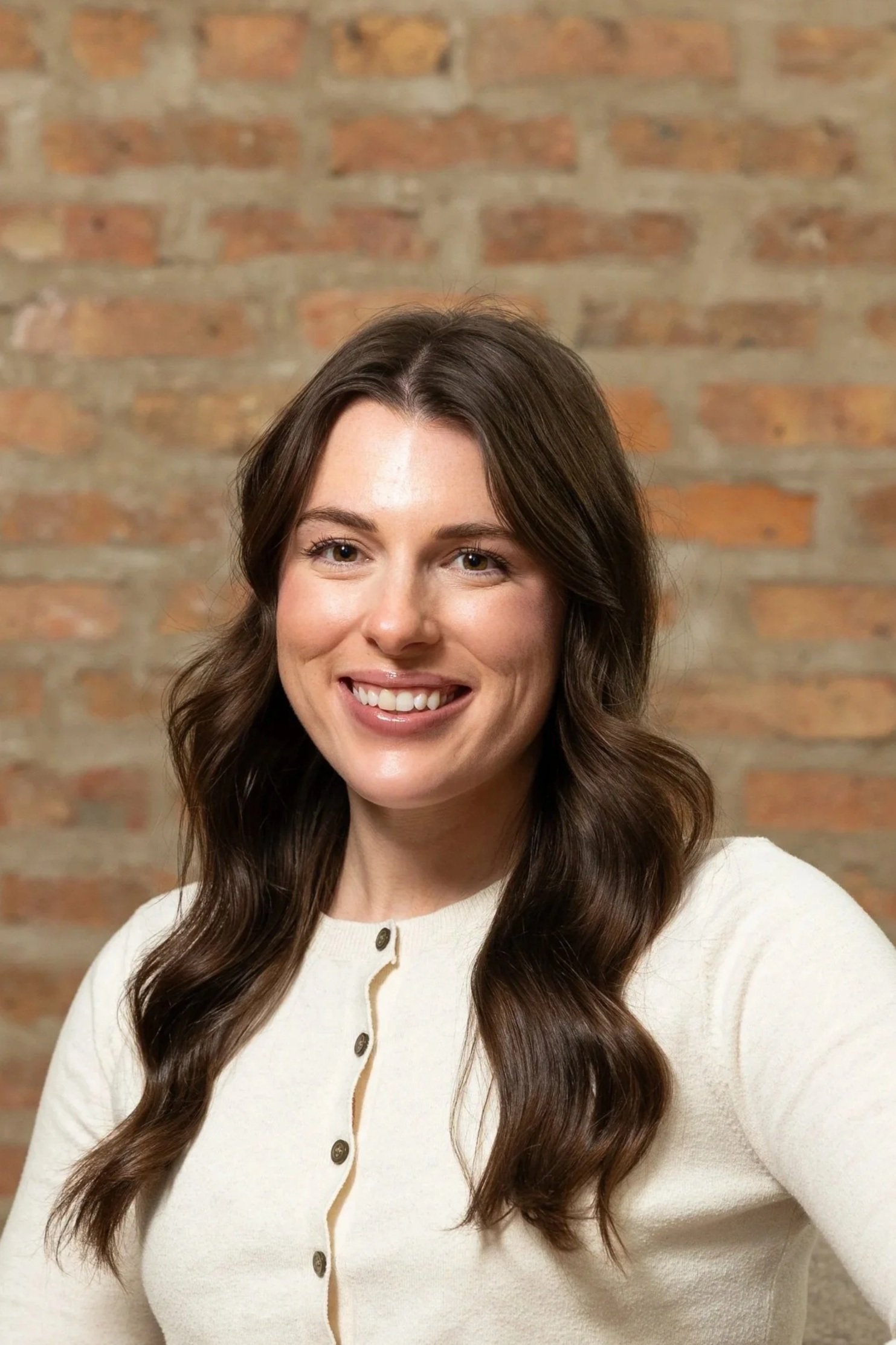 A smiling woman with long, wavy brown hair wearing a cream-colored cardigan, standing in front of a brick wall.